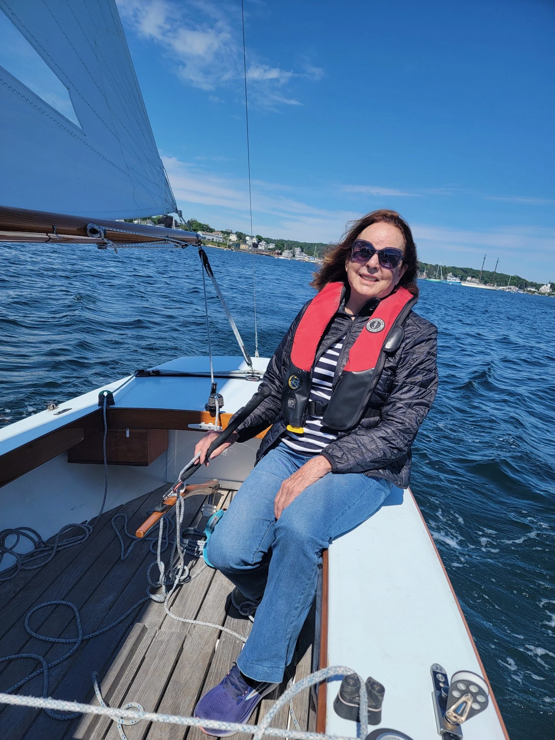Woman with sunglasses and a red life jacket sitting on a boat in the water, smiling, with a scenic shoreline in the background under a clear blue sky.