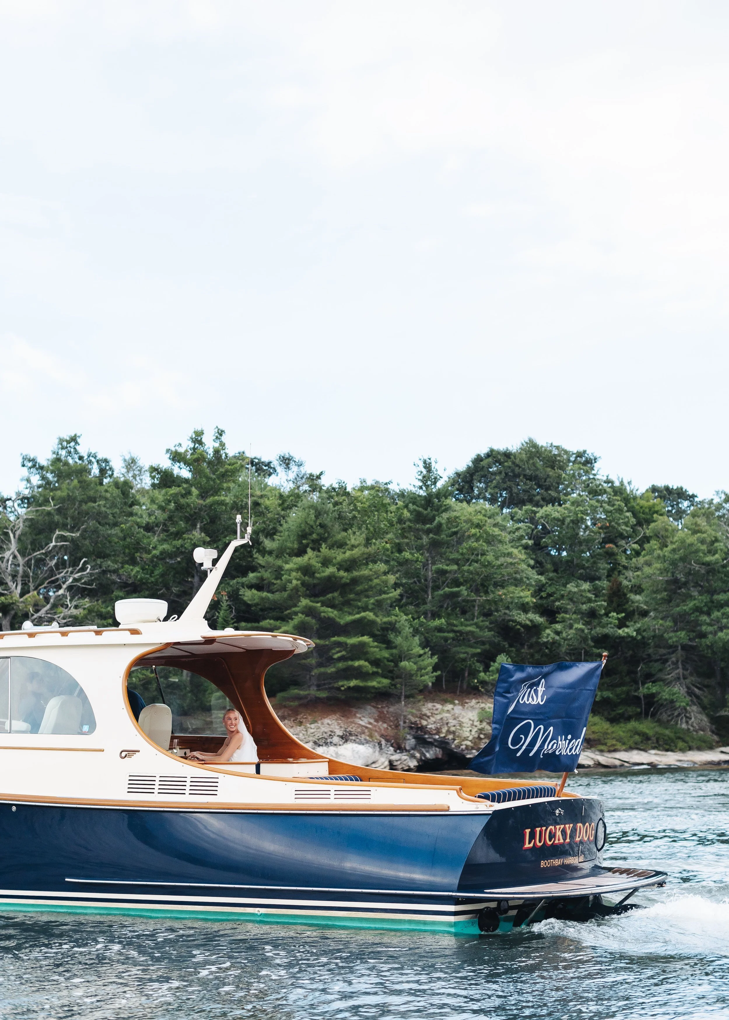 A boat named 'Lucky Dog' with a 'Just Married' banner, cruising on a river near a forested shore, with a woman smiling in the window.