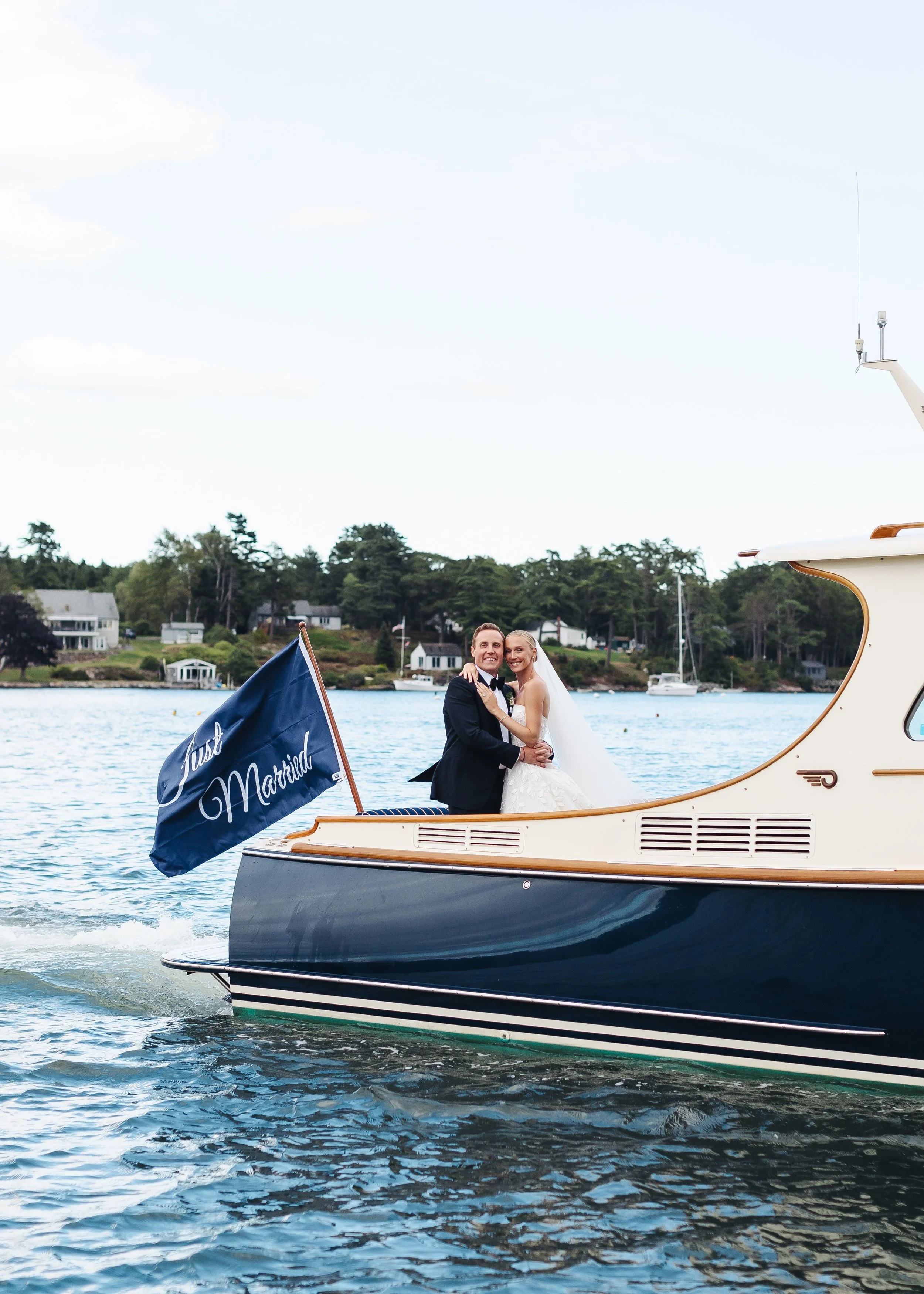 a newlywed couple embracing on a boat with a 'Just Married' flag on a body of water, with houses and trees in the background.