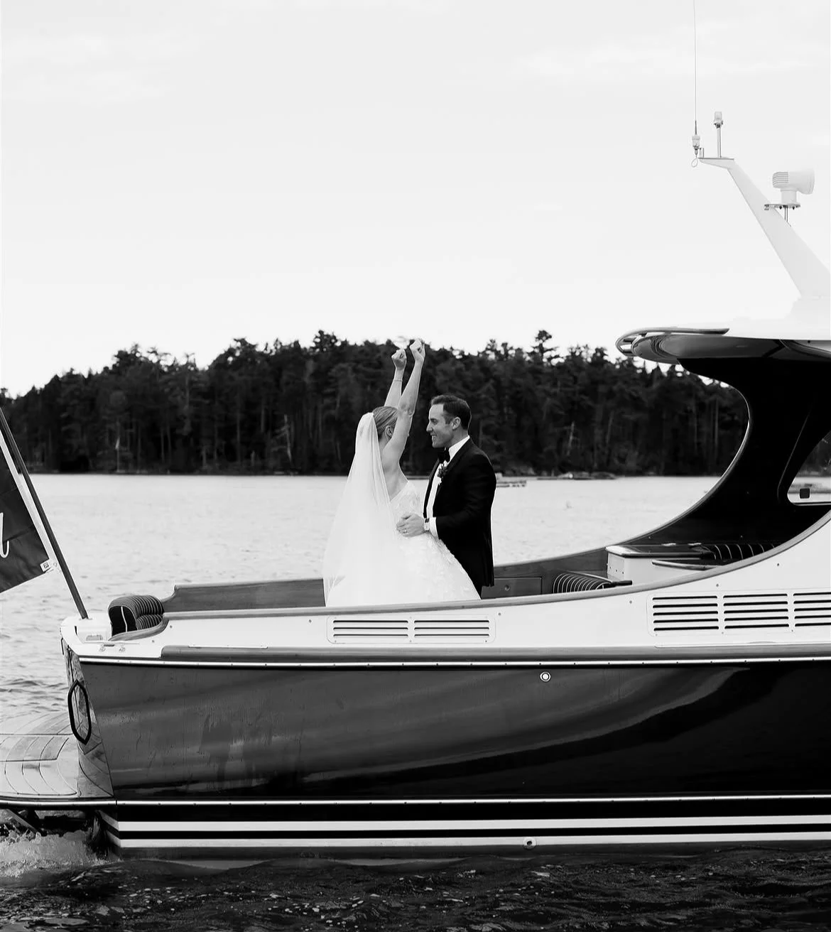 A bride and groom dancing on a yacht with water and forest in the background, captured in black and white.