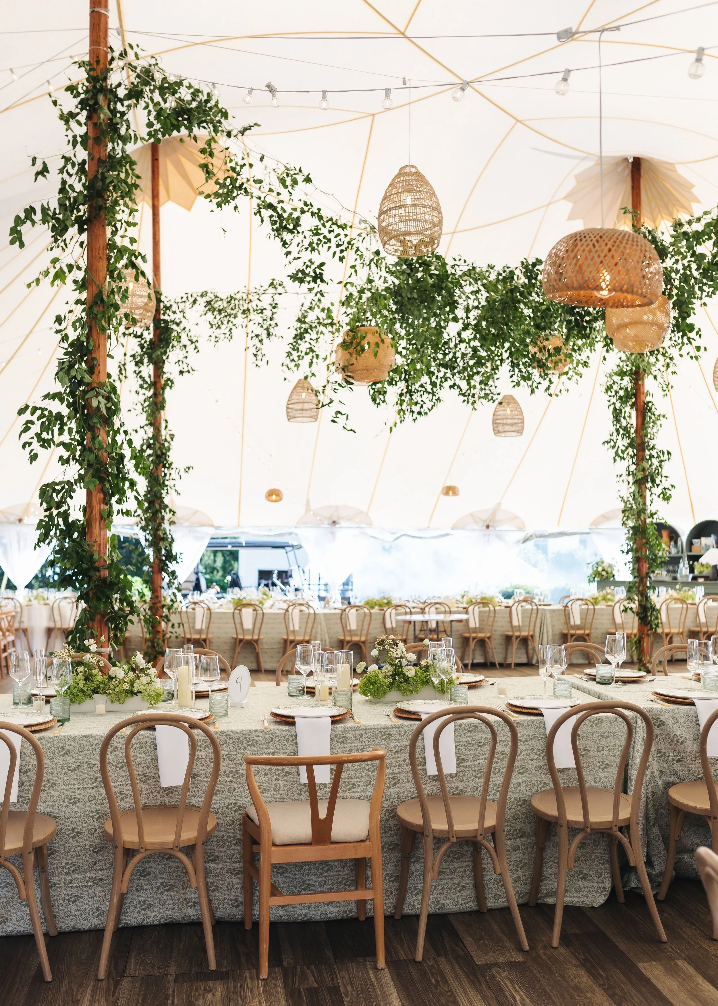 A decorated event tent with hanging wicker lamps and greenery, set for a formal meal with tables, chairs, and table settings including floral centerpieces and candles.