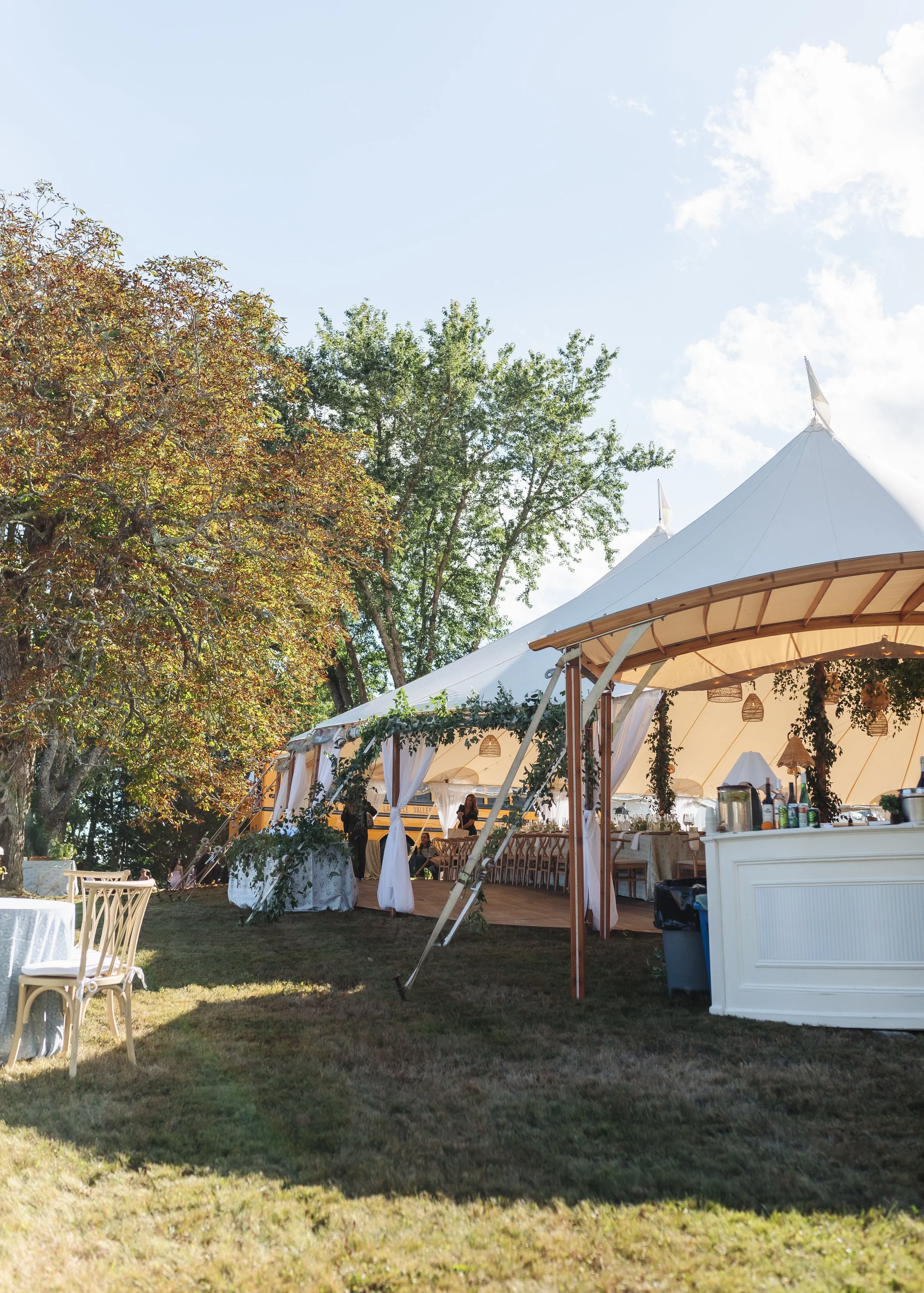 Outdoor event setup with a large white canopy tent, decorated with trees and string lights, under a clear blue sky.