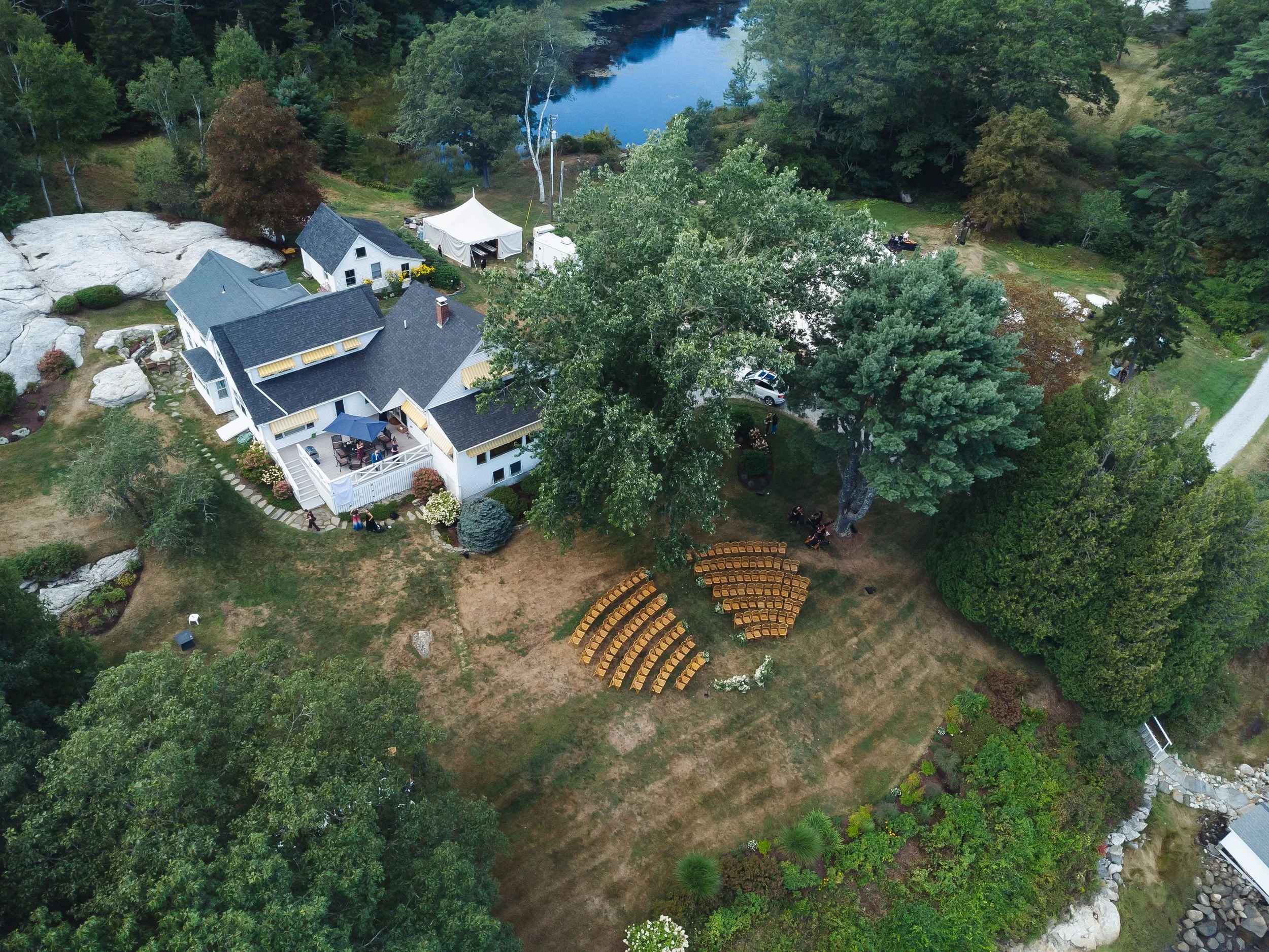 Aerial view of a waterfront house with a dark roof, a small white tent, and multiple trees surrounding a lawn with yellow chairs arranged in semi-circular rows. There is a pond in the background and a gravel path leading to the house.