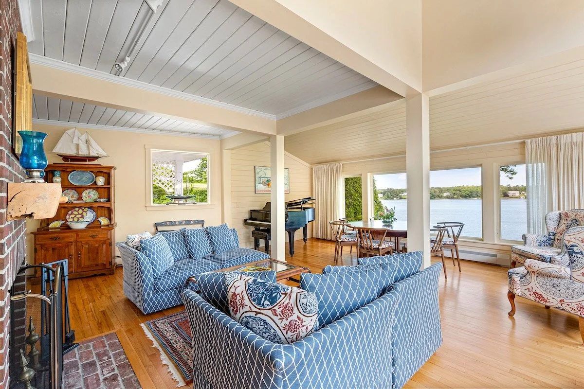 Living room with blue patterned sofas, a wooden dining table, large windows overlooking a harbor, a grand piano, and a nautical décor theme.