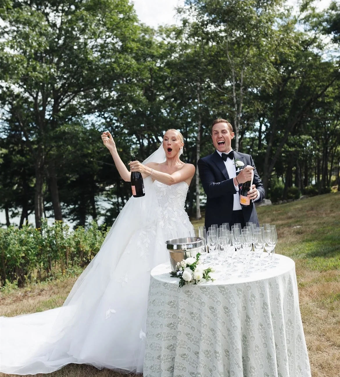 A bride and groom celebrating outdoors on their wedding day, opening bottles of champagne over a table with glasses.