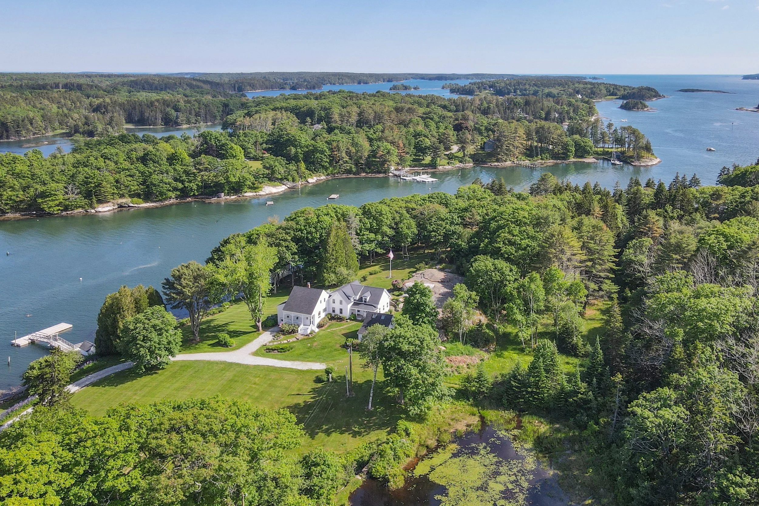 Aerial view of a coastal area with lush green trees, houses near the water, and boats docked along the shoreline on a sunny day.