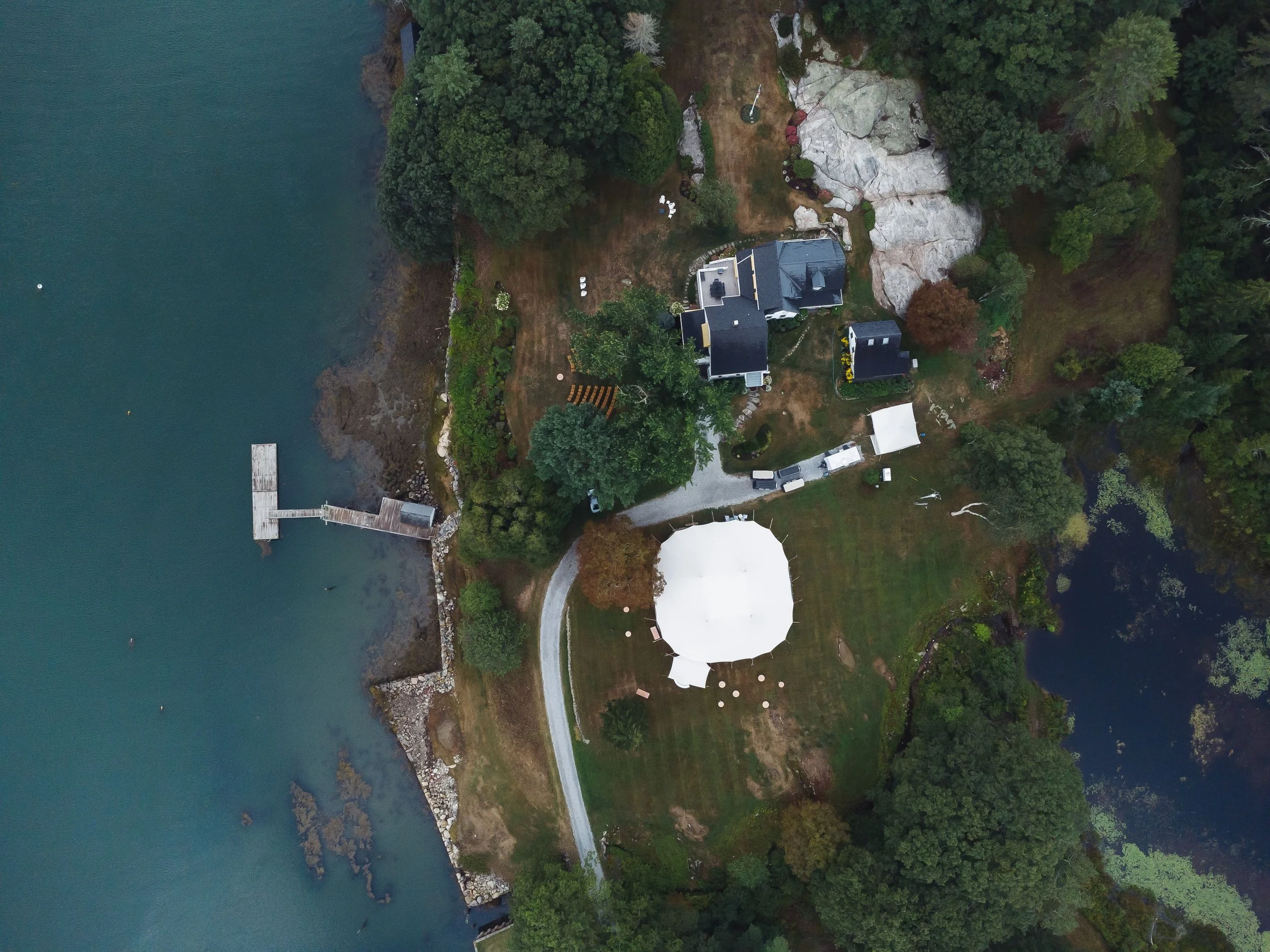 An aerial view of a house on a harbor surrounded by trees and a rocky landscape, with a dock extending into the water.
