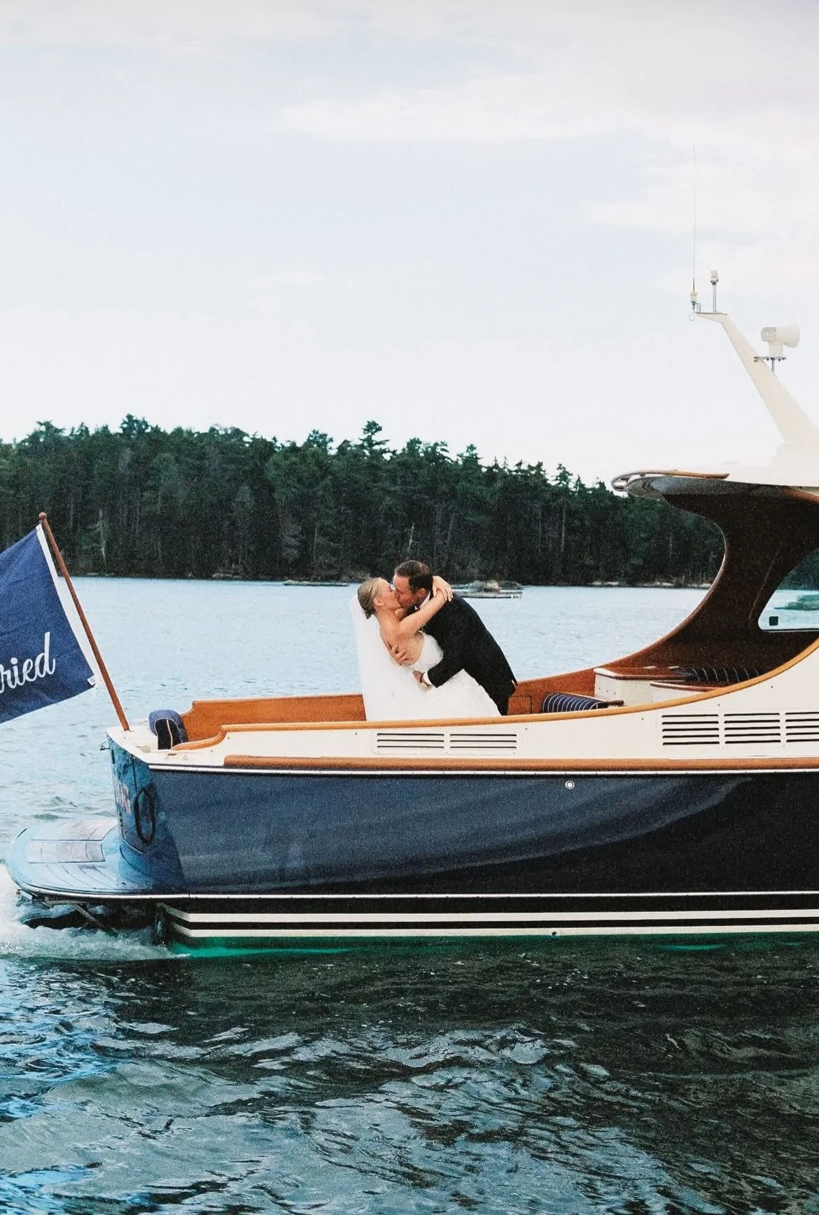 A bride and groom sharing a kiss on a boat on the water, with trees in the background.