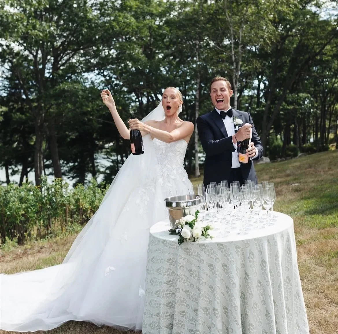 Bride and groom celebrating with champagne at outdoor wedding reception, table with glasses and flowers, trees in background.