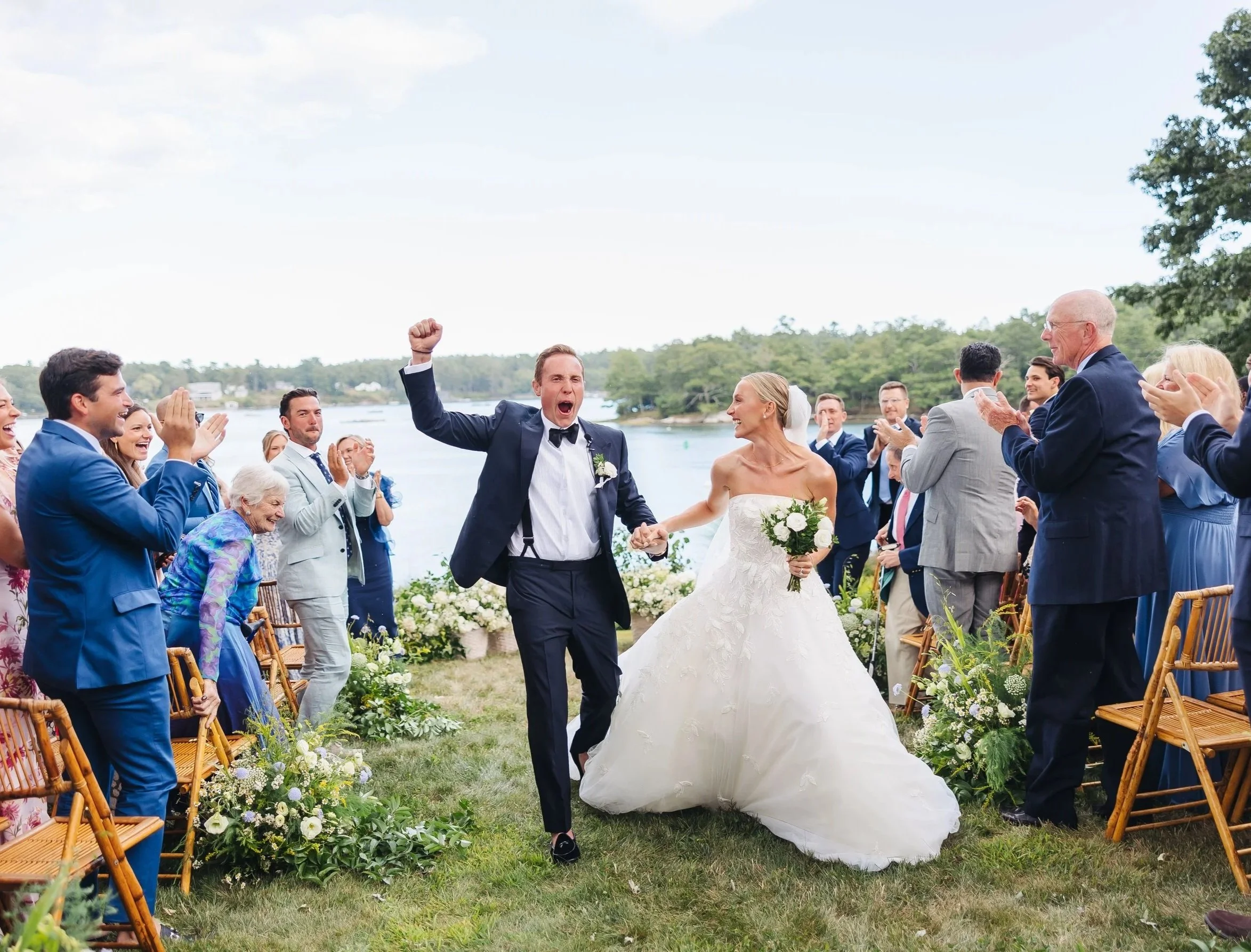 A wedding celebration outdoors by a harbor, with a bride and groom walking down an aisle surrounded by friends and family clapping and cheering.