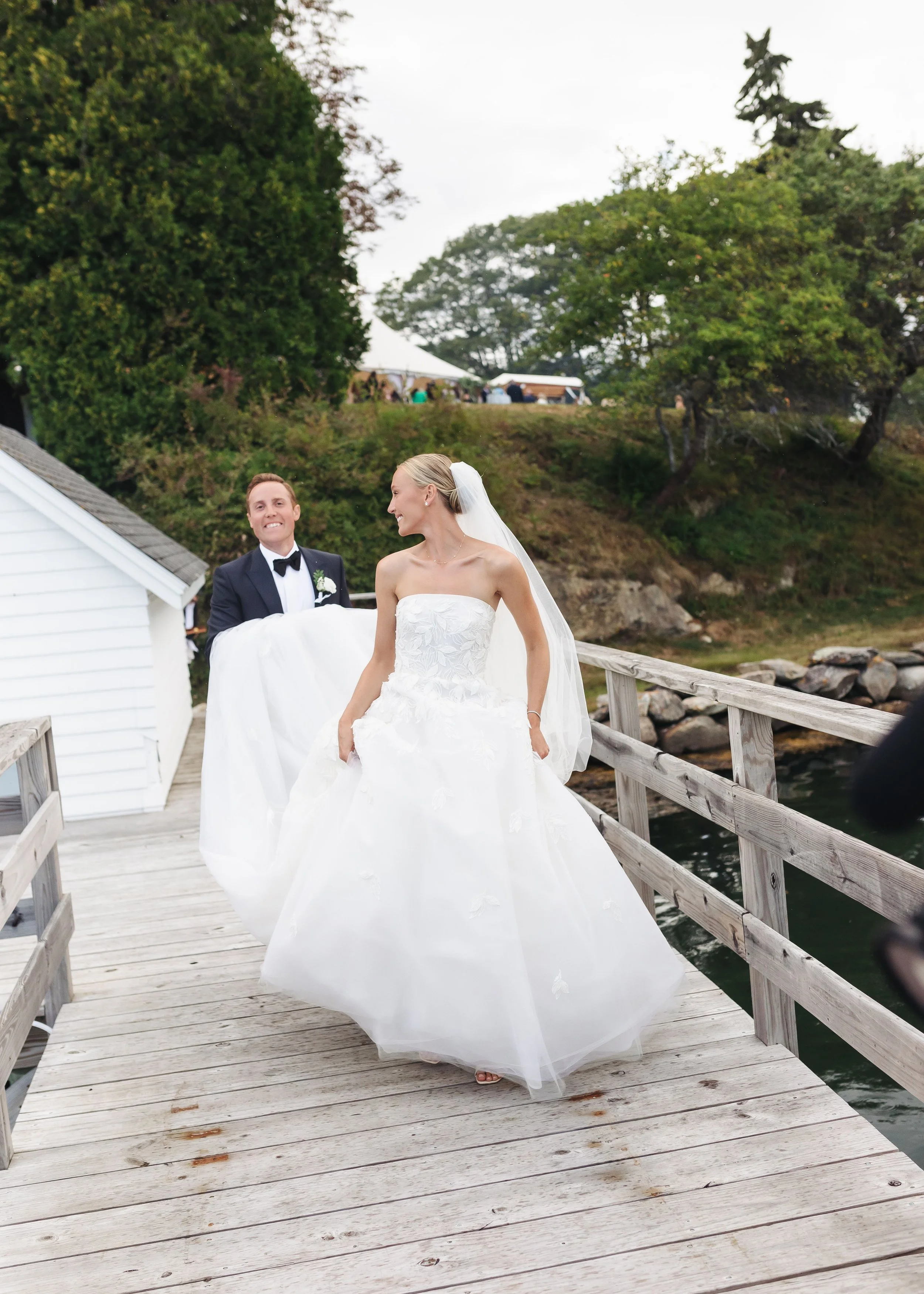 A bride in a strapless white wedding gown and veil, walking on a wooden dock while a groom in a black tuxedo and bow tie walks behind her, at an outdoor waterfront wedding venue with green trees and a rocky shoreline in the background.
