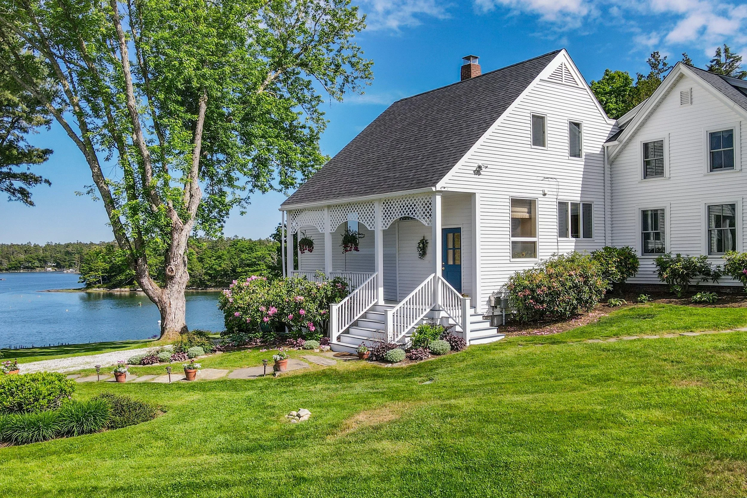 A white, two-story house with a steep roof and a front porch with decorative trim, situated near a body of water and surrounded by green lawns and trees, under a bright blue sky.