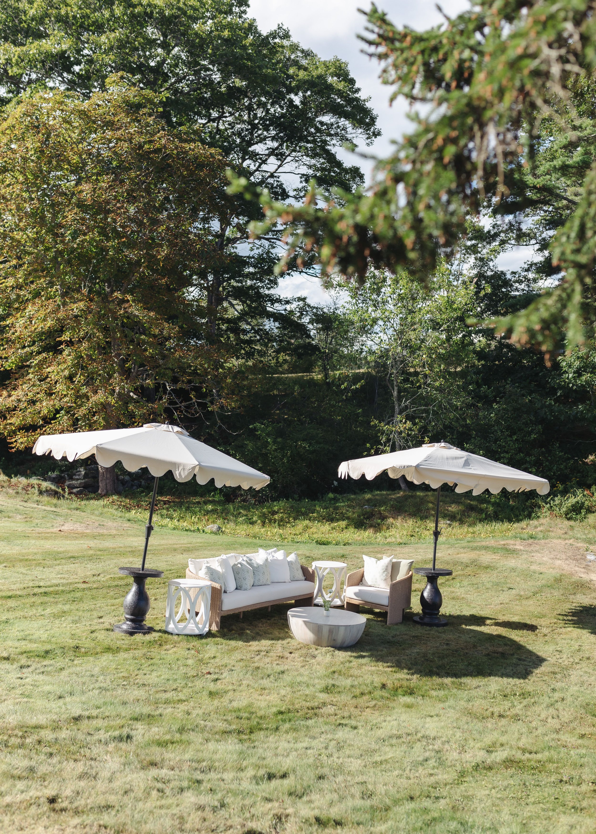 Outdoor seating area on a grassy lawn with white umbrellas, white sofa, armchair, small coffee tables, and decorative pillows surrounded by trees and greenery.