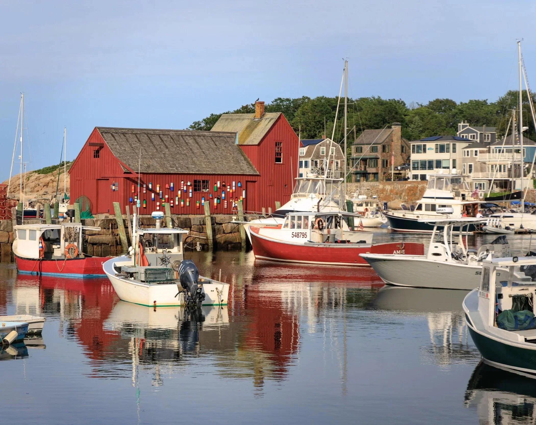 A marina with various boats docked in the water, a red building decorated with colorful buoys, and residential houses with greenery in the background under a blue sky.