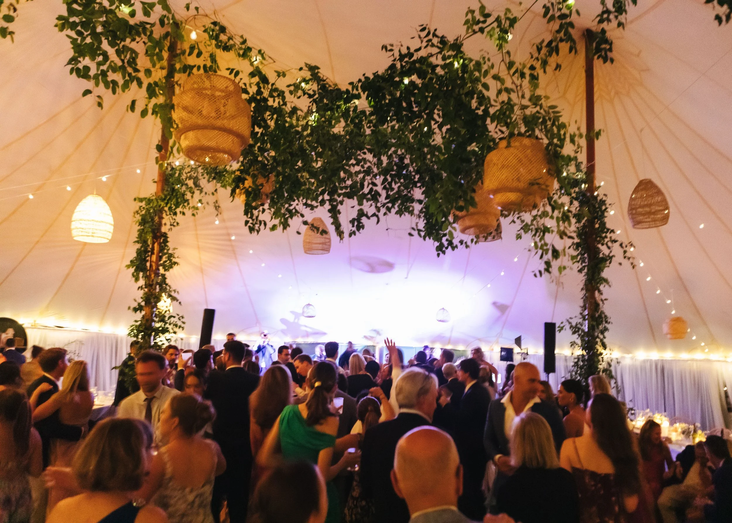 A crowded indoor event with a large group of people dancing and socializing underneath a decorated ceiling with hanging lanterns and greenery.