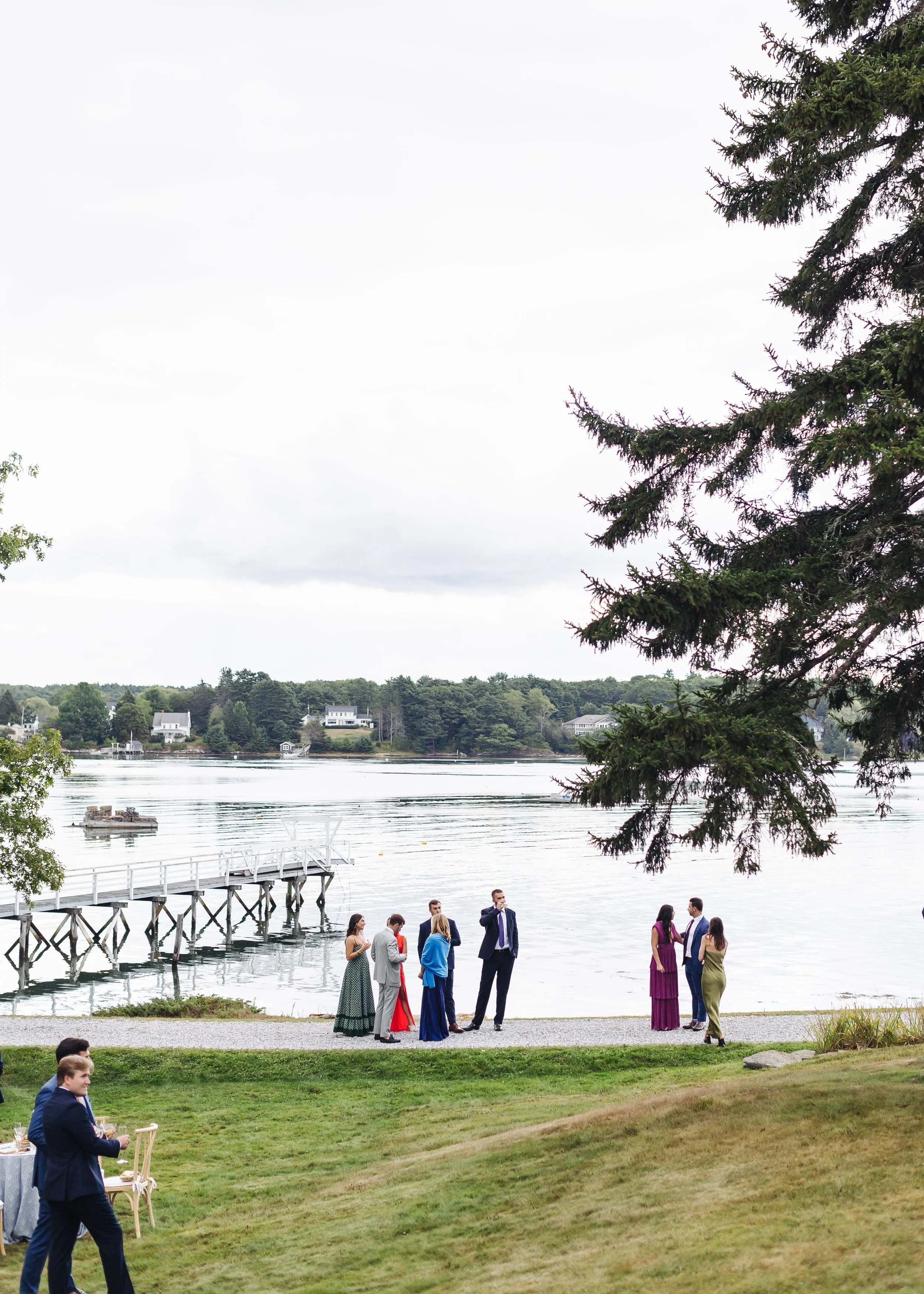 A group of people dressed in formal attire standing and socializing by a waterfront with houses and trees in the background. Some are talking near a dock, and there is a man in the foreground walking near a table with drinks.