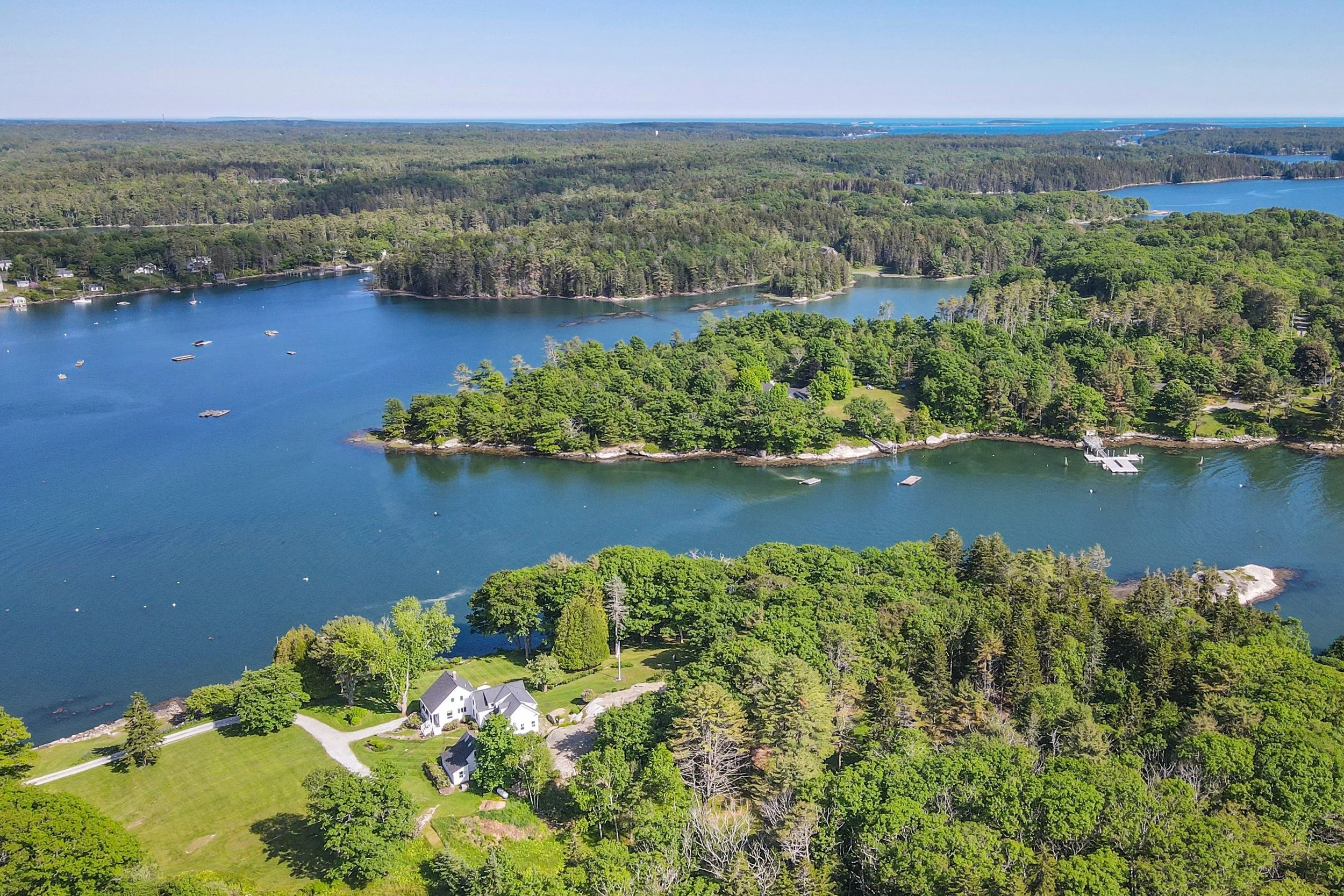 Aerial view of a coastal landscape with green forests, a harbor, and houses with docks, under a clear blue sky.