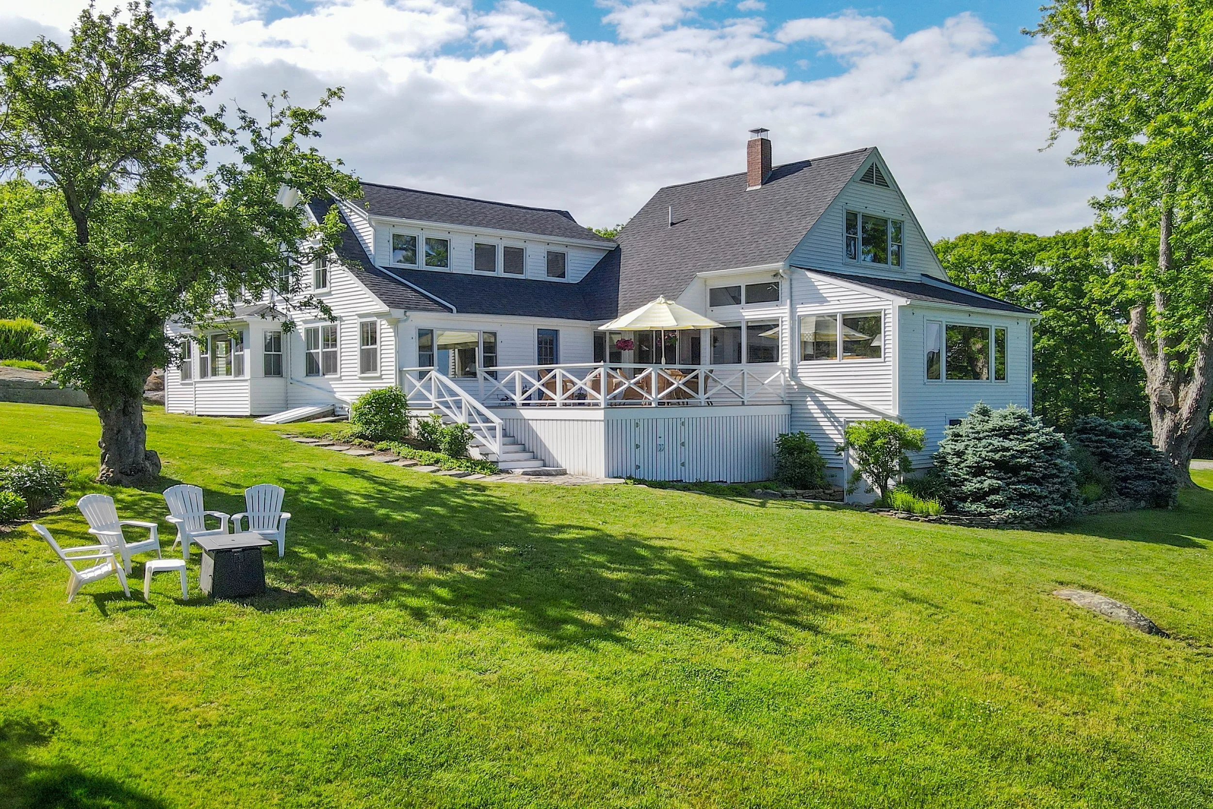White house with multiple windows and a large deck, surrounded by green trees and a well-maintained lawn with outdoor chairs.