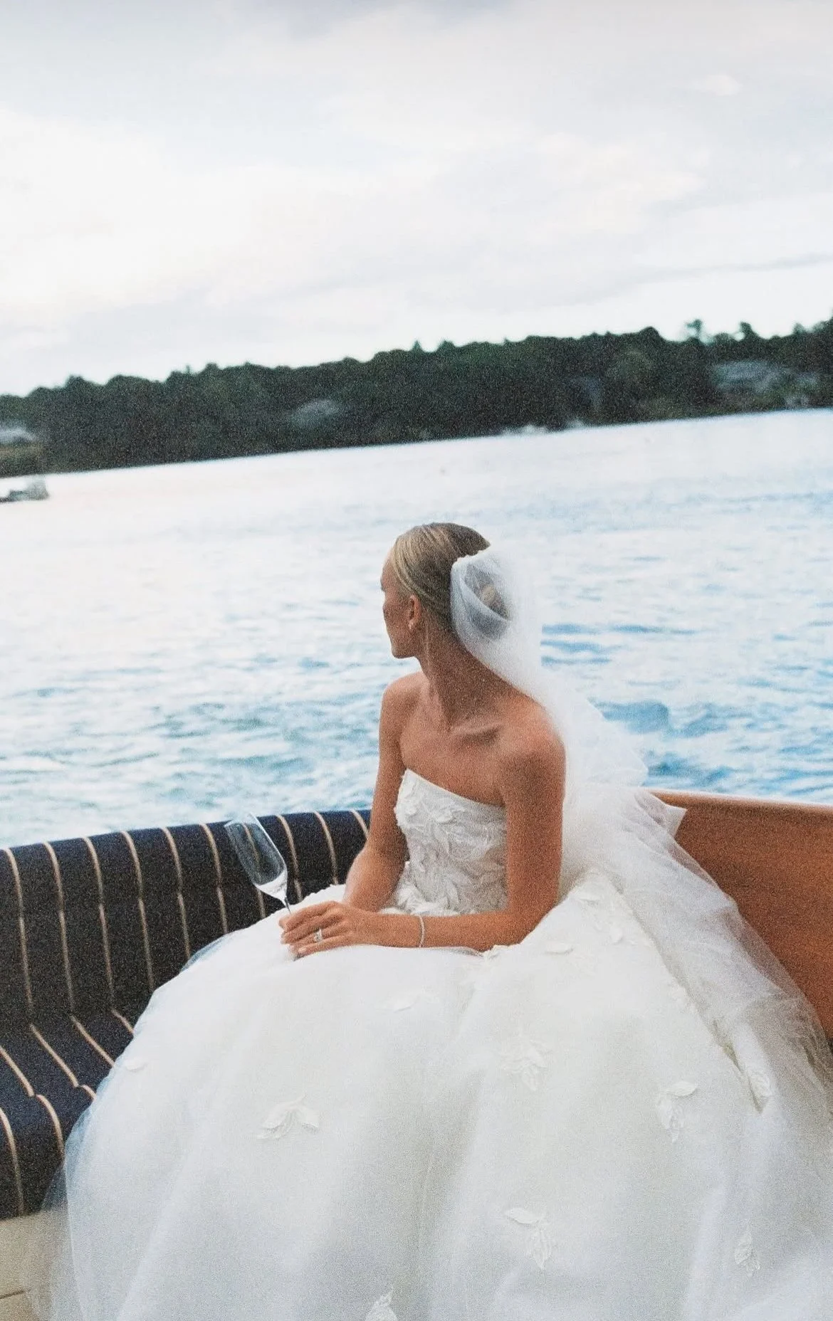 A bride sitting on a boat railing with a wedding dress and veil, holding a glass of wine, looking away from the camera, on a body of water with trees and houses in the background.