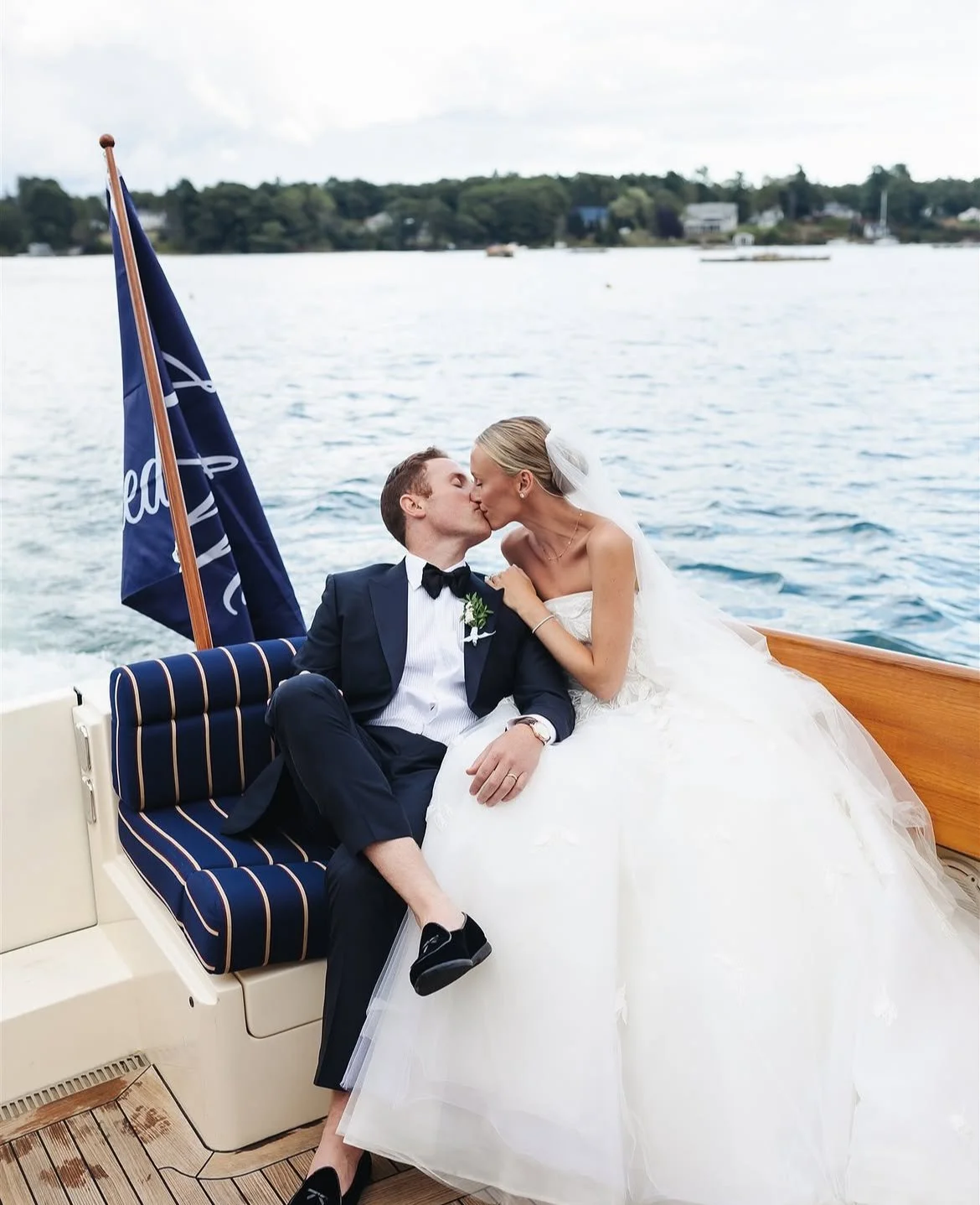 A newlywed couple in wedding attire sharing a kiss on a boat, with water and trees in the background.