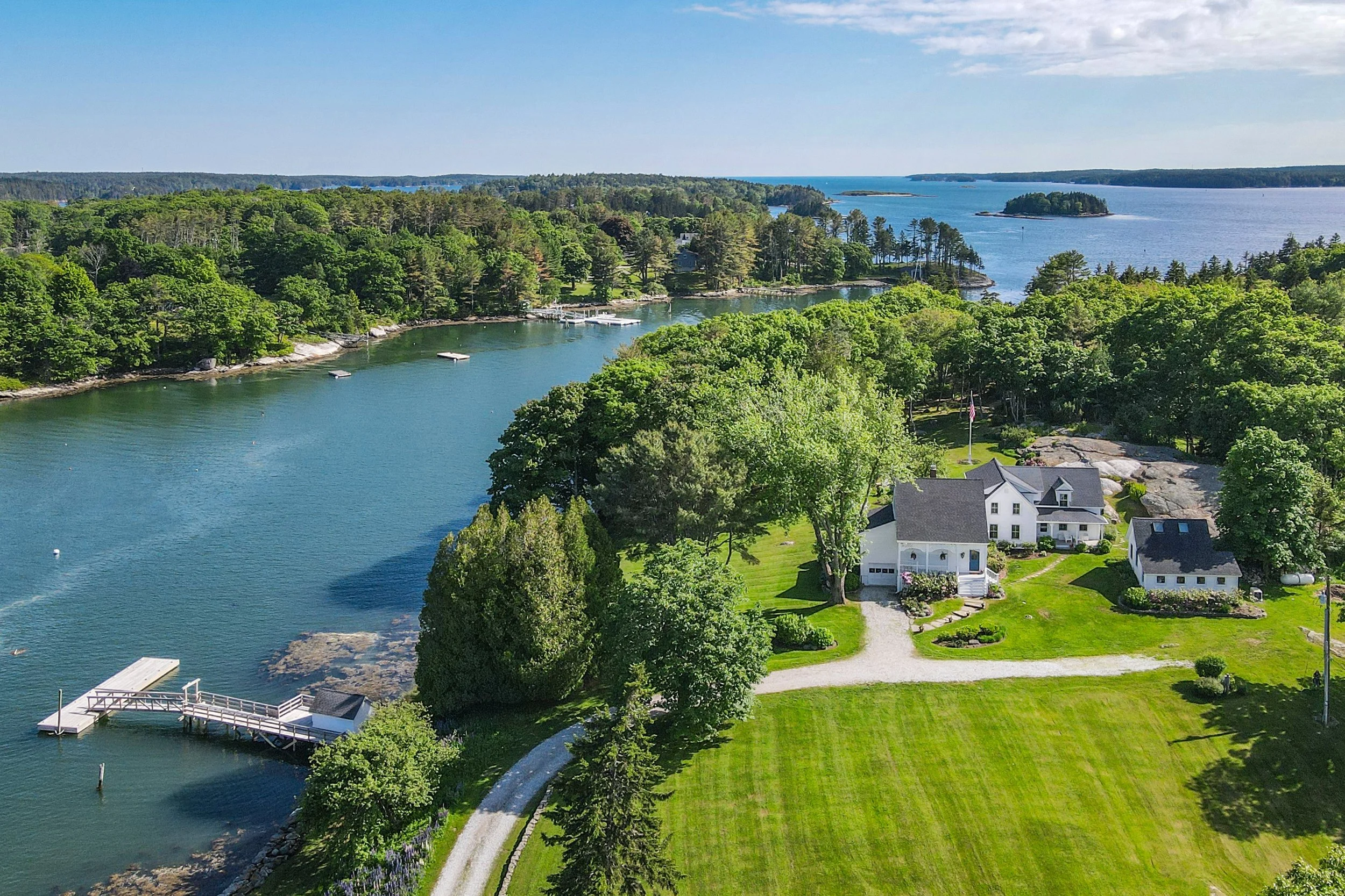 Aerial view of a waterfront estate with a white house, a grassy lawn, a dock, and a forested shoreline extending to the ocean in the background.