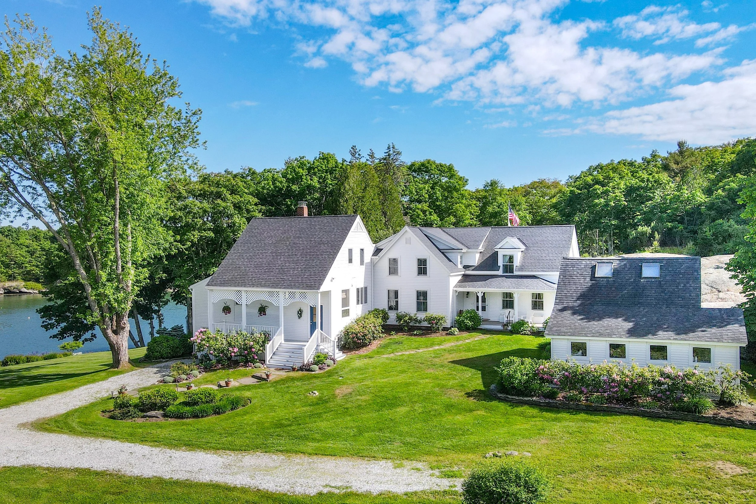 A white house with a porch, surrounded by a manicured lawn, colorful flowers, and lush green trees, with a view of a harbor and a blue sky with scattered clouds.