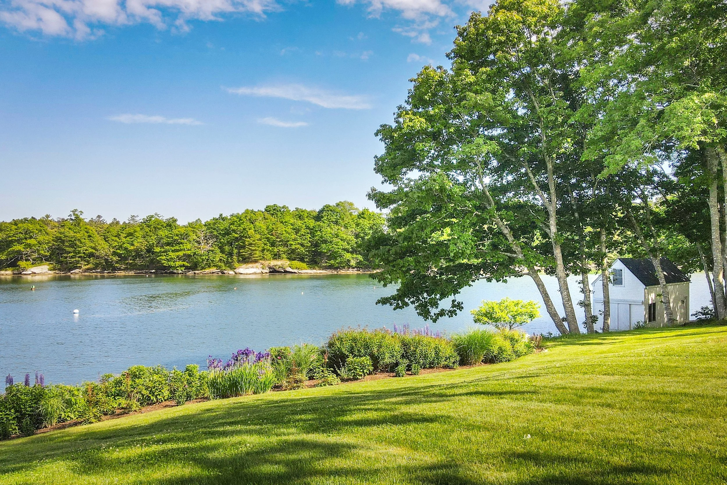 A peaceful harbor scene with a grassy yard, colorful flowers, large green trees, calm water, and a small white boathouse house with a black roof near the trees.