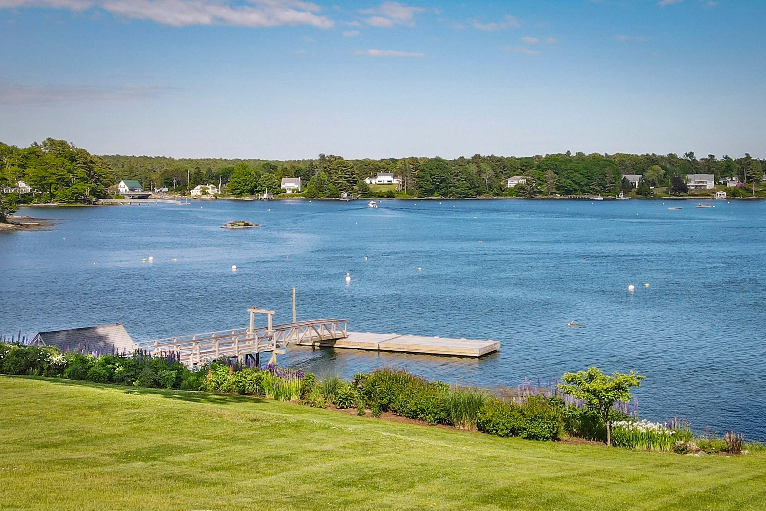 Scenic view of a harbor with houses and trees along the shoreline, a dock extending into the water, and a clear sky.