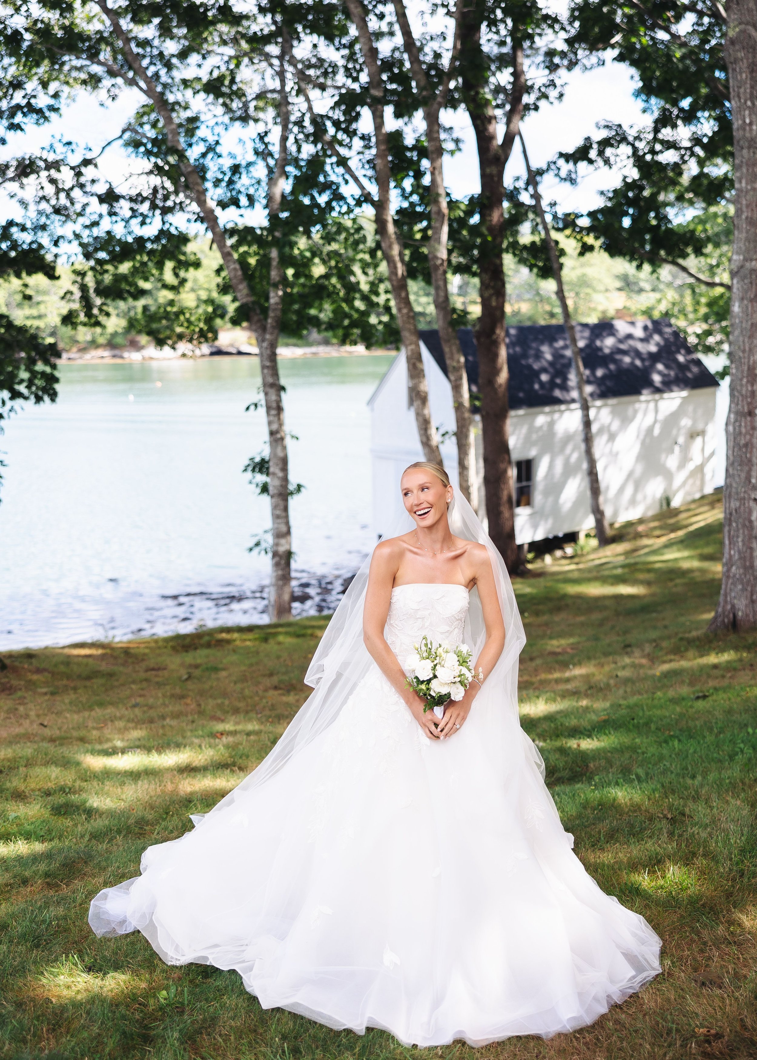 A bride in a white wedding dress holding a bouquet of white flowers stands outdoors on grass near a body of water, with trees and a white building with a black roof in the background. She is smiling and looking to the side.