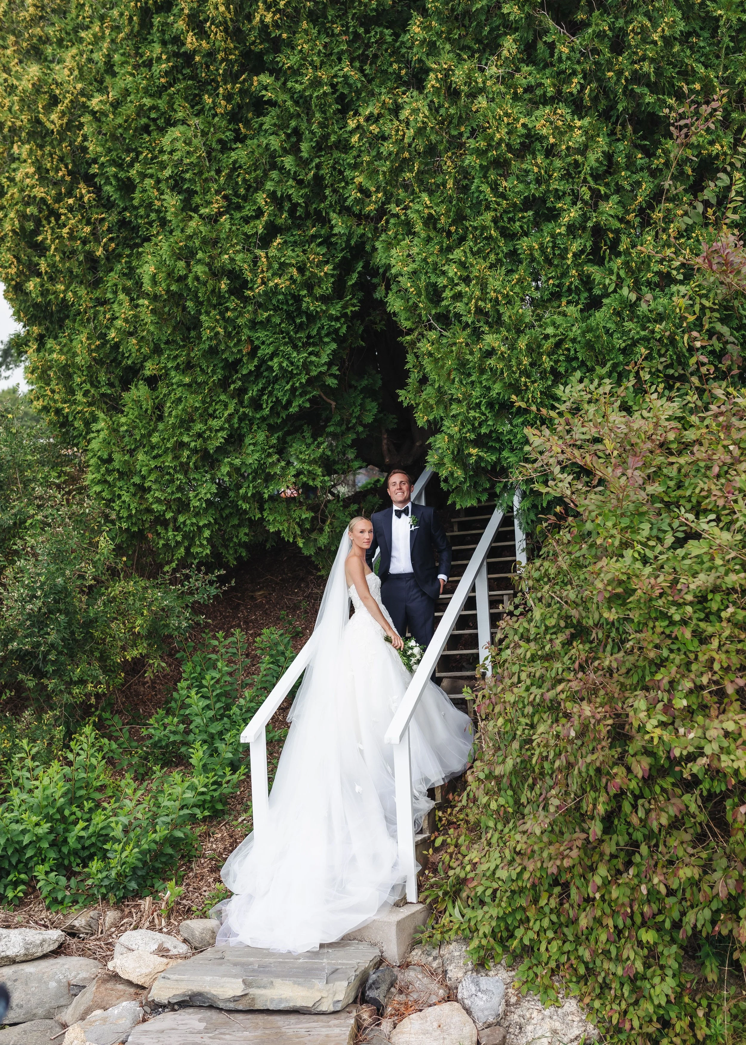 A bride and groom in wedding attire standing on an outdoor staircase surrounded by lush green foliage. The bride wears a white wedding dress with a long train and veil, and the groom wears a black tuxedo with a bow tie. They are smiling and looking t