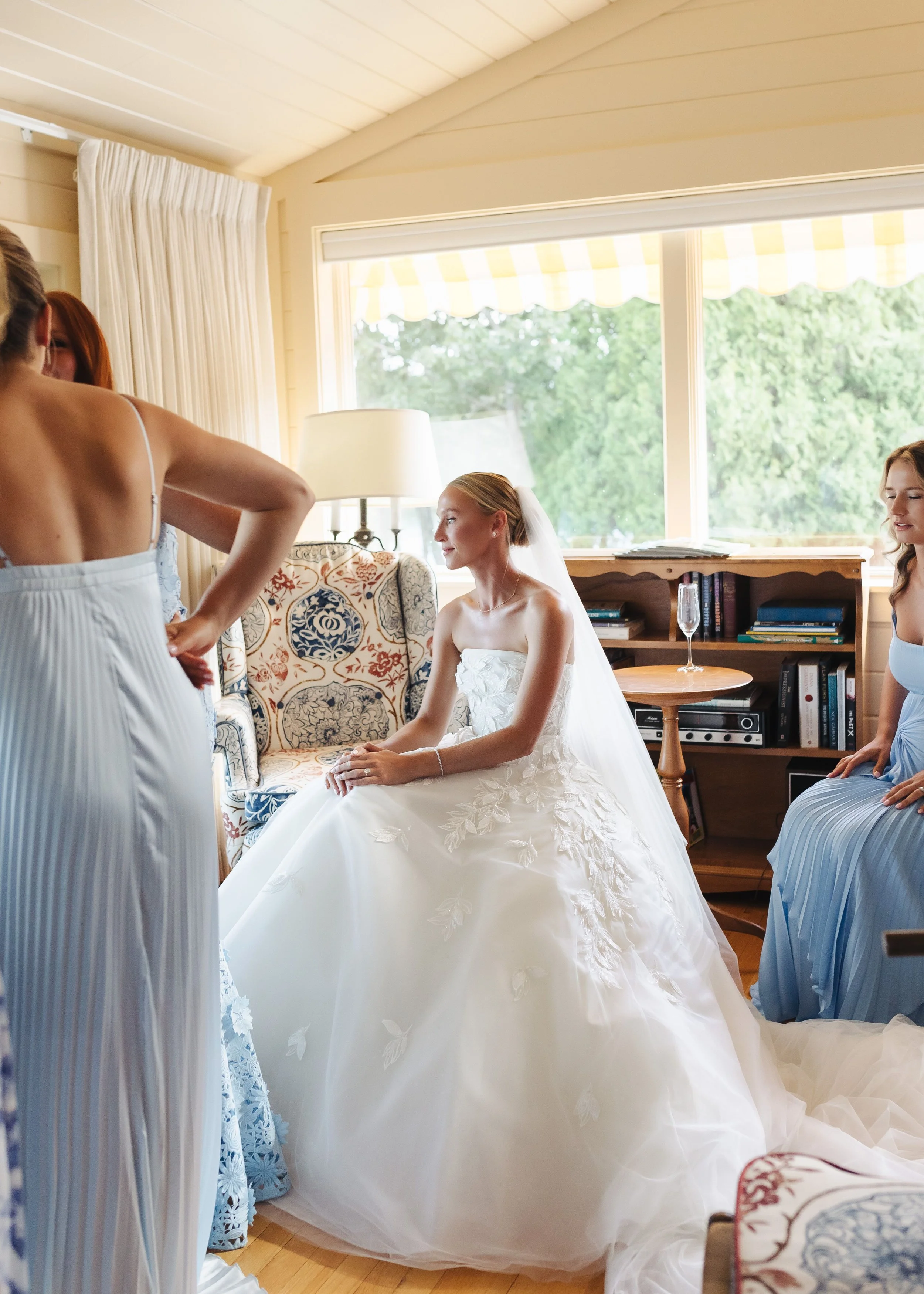 Bride sitting on a floral armchair wearing a wedding dress with embroidered leaves, bride smiling and looking to the side, bridesmaids in light blue dresses assisting her, large window with yellow-and-white striped awning outside, bookshelf with book