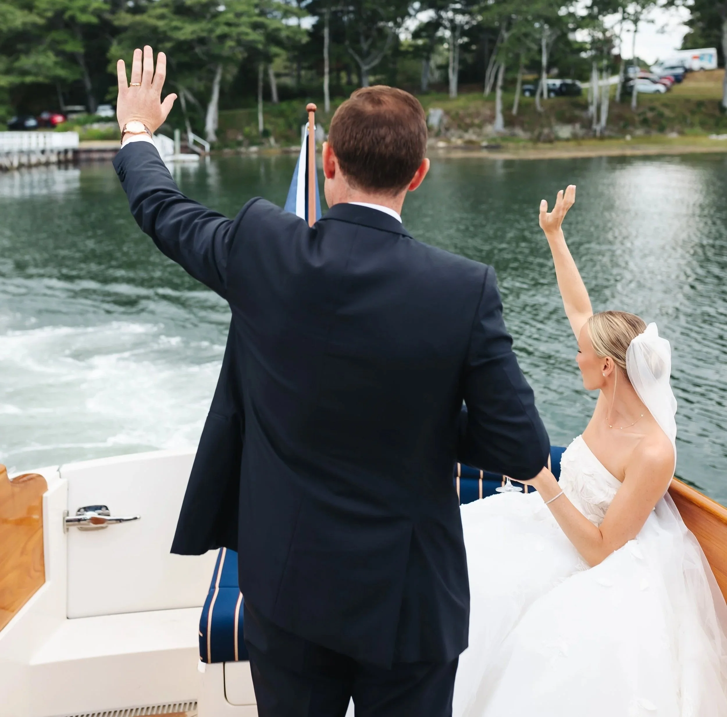 A man in a tuxedo and a woman in a wedding dress on a boat, with her hand on his shoulder, enjoying a scenic waterway surrounded by trees and parked cars.