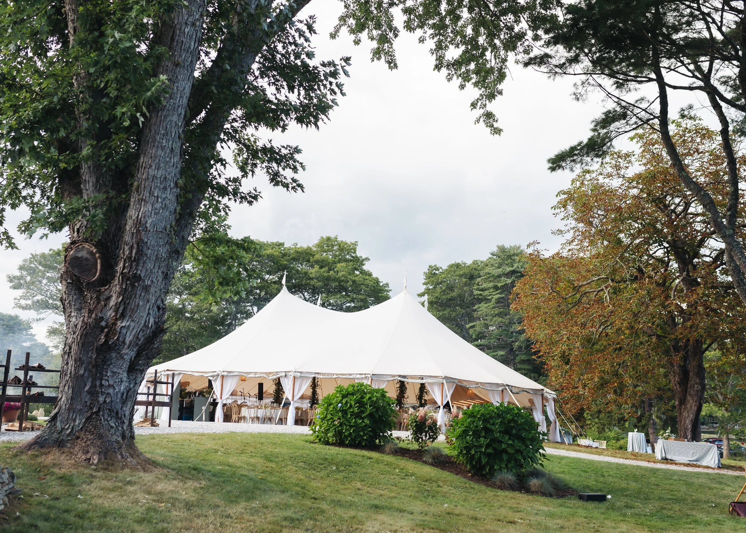 A large white event tent set up outdoors in a park-like area with large trees and green grass, suggesting a gathering or celebration.