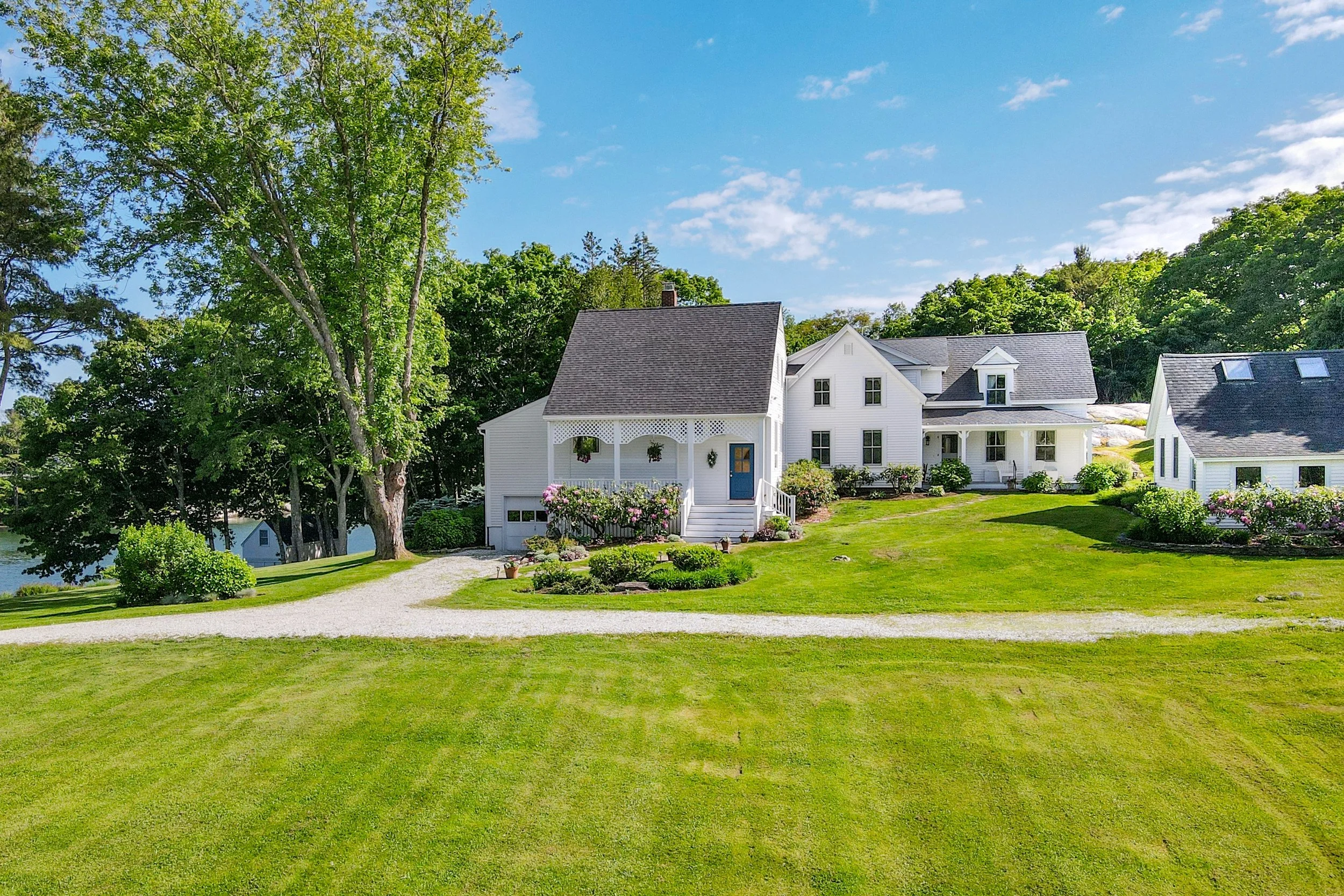 A white house with a porch, surrounded by green trees and maintained lawn, under a blue sky with a few clouds.