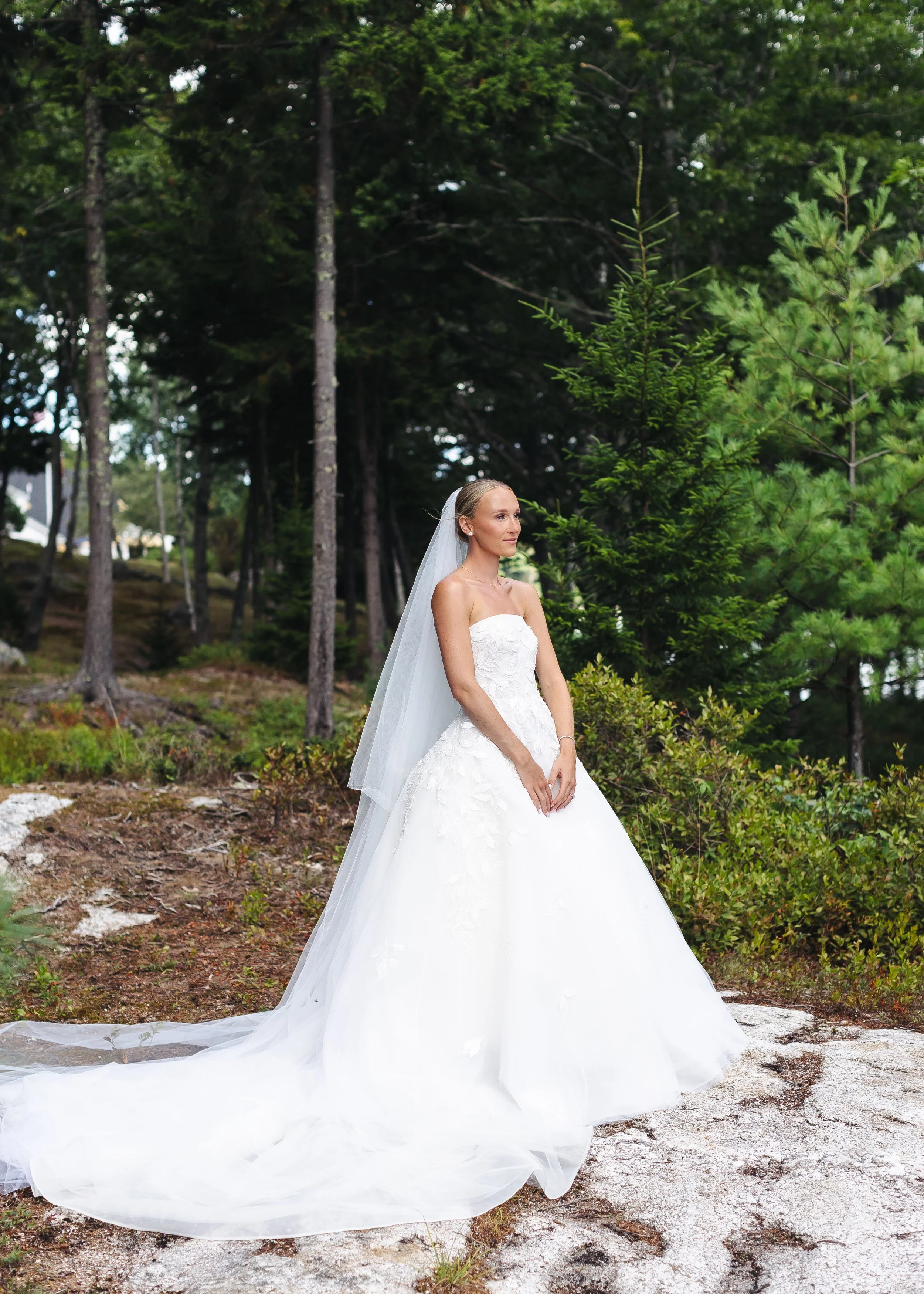A bride standing outdoors in a white wedding dress with a long train, surrounded by trees and greenery.