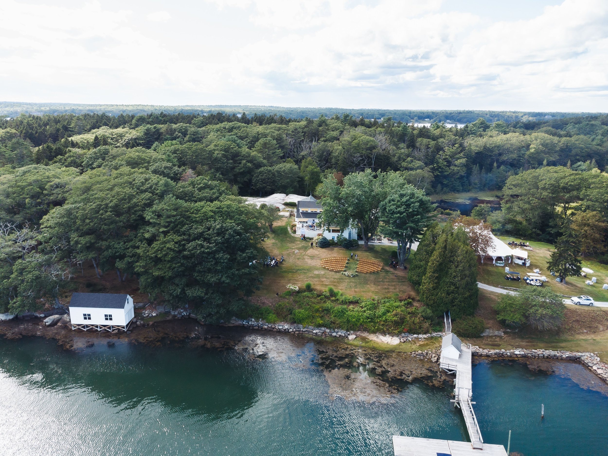 Aerial view of a wooded landscape with a small white house near the water, surrounded by trees and a dock extending into a river.