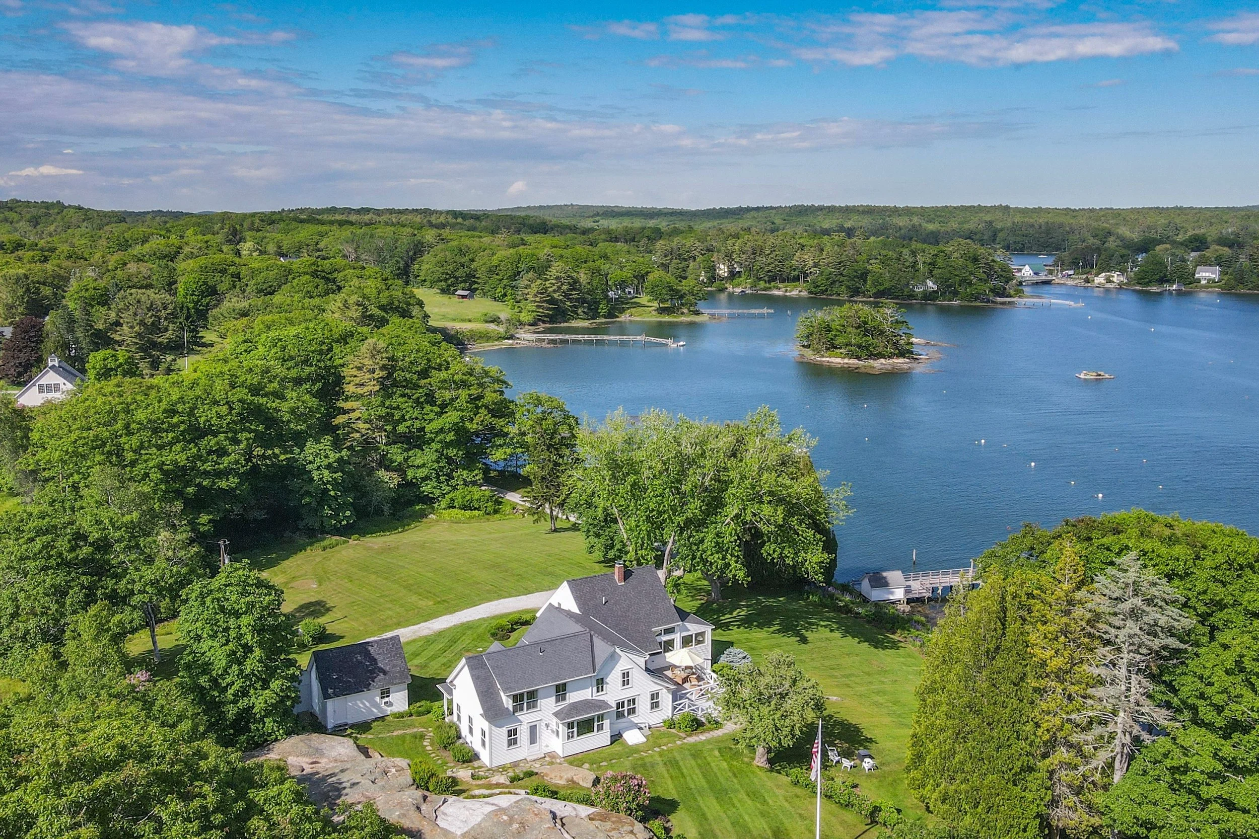 Aerial view of a waterfront estate with greenery, a house, docks, and boats on the water under a partly cloudy sky.