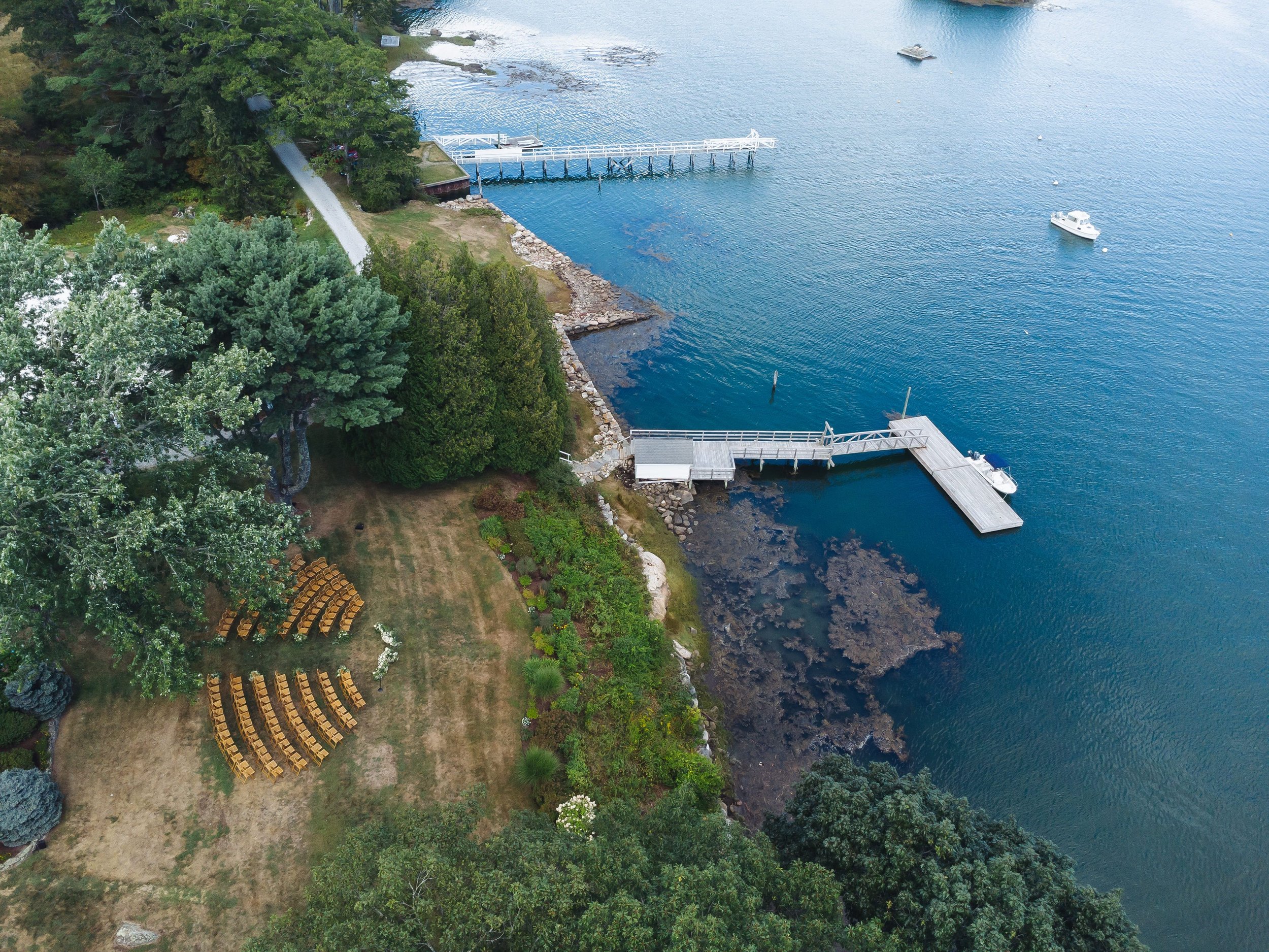Aerial view of a waterfront landscape with a wooden dock extending into the water, surrounded by lush green trees and grass. There are a few boats on the water and a small rocky shoreline.