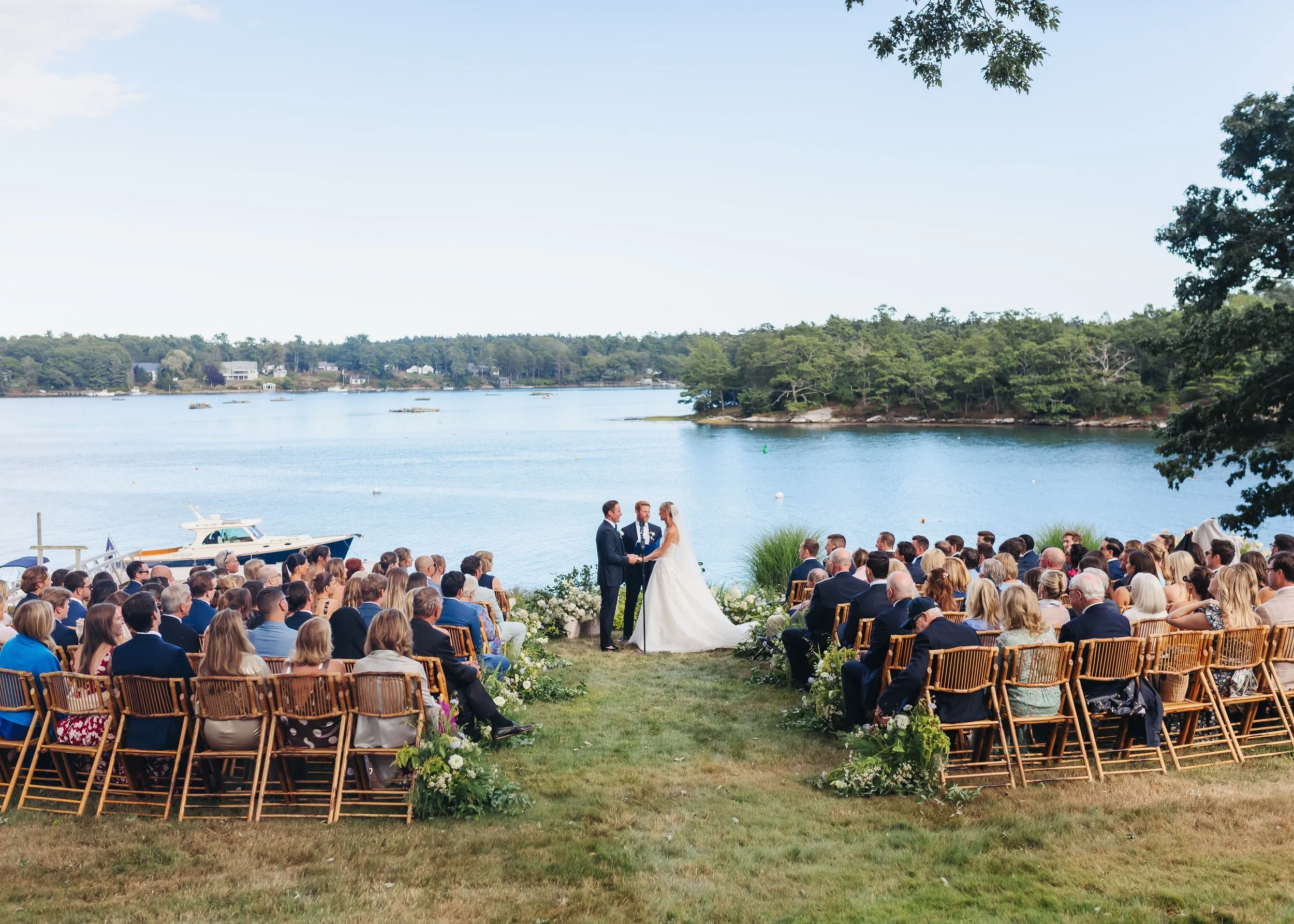 A large outdoor wedding ceremony with a bride and groom standing under a tree, facing a seated audience near a body of water, with trees and houses in the background.