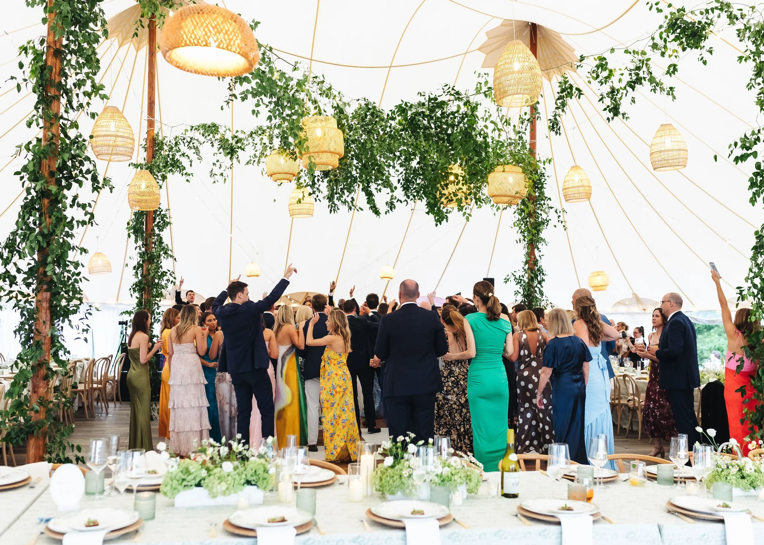 A wedding reception under a tent with hanging lights, green foliage, and a crowd of guests in formal attire dancing and mingling.