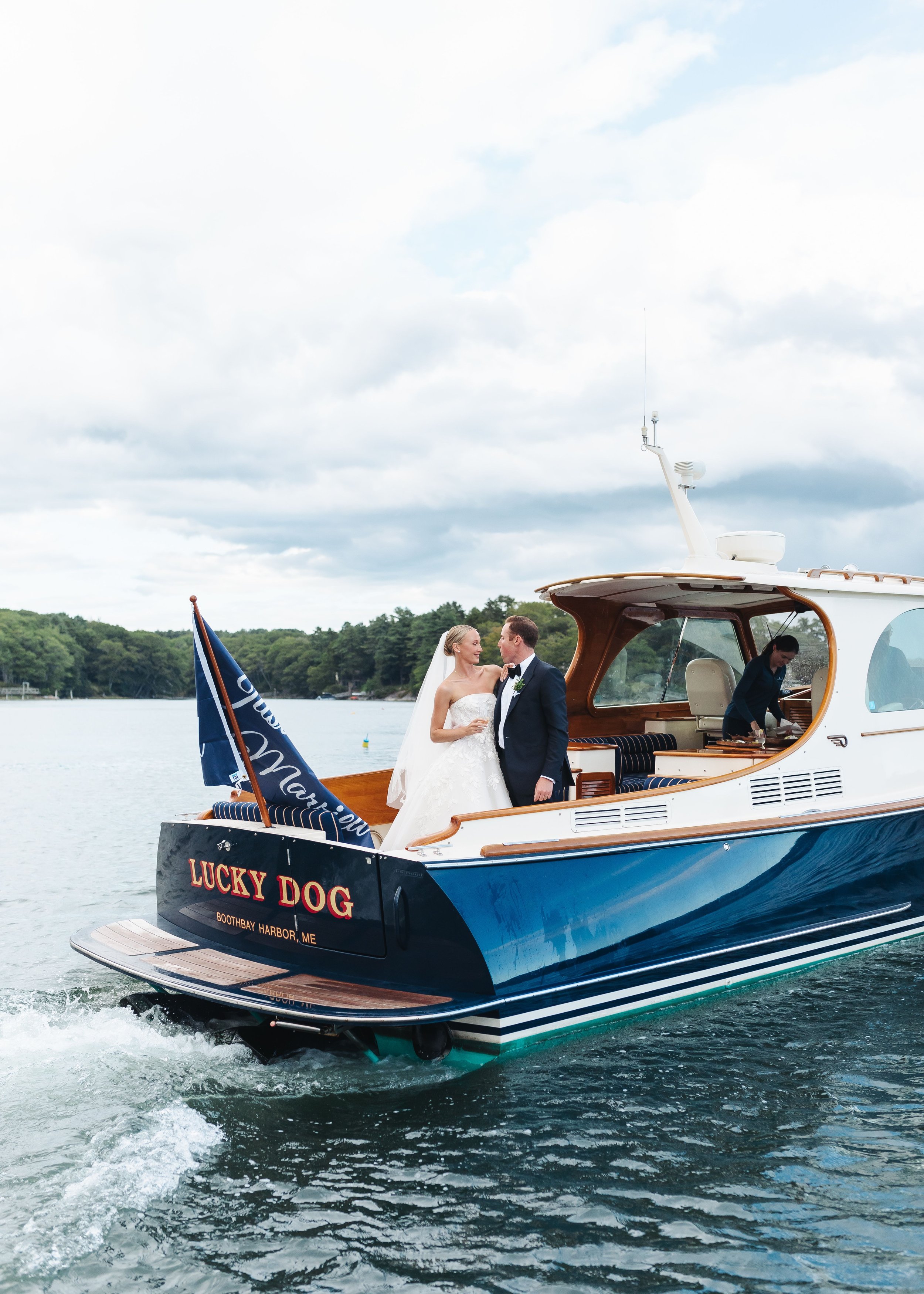 A couple in wedding attire standing on a boat named 'Lucky Dog' on the water, with a woman cooking in the background, and a flag on the boat, under a partly cloudy sky.