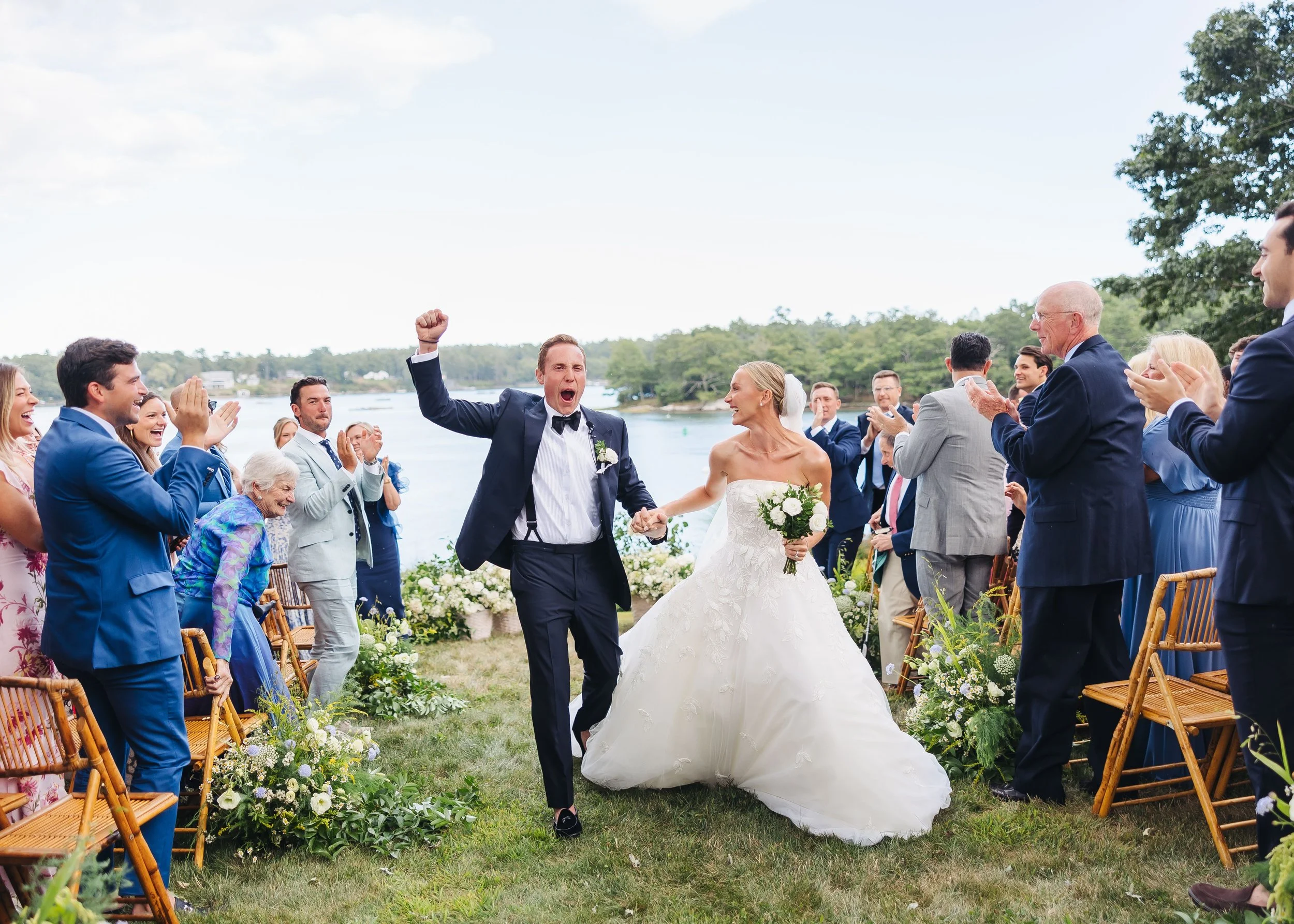 A bride and groom celebrating during their outdoor wedding, surrounded by friends and family, with a scenic water and tree background.