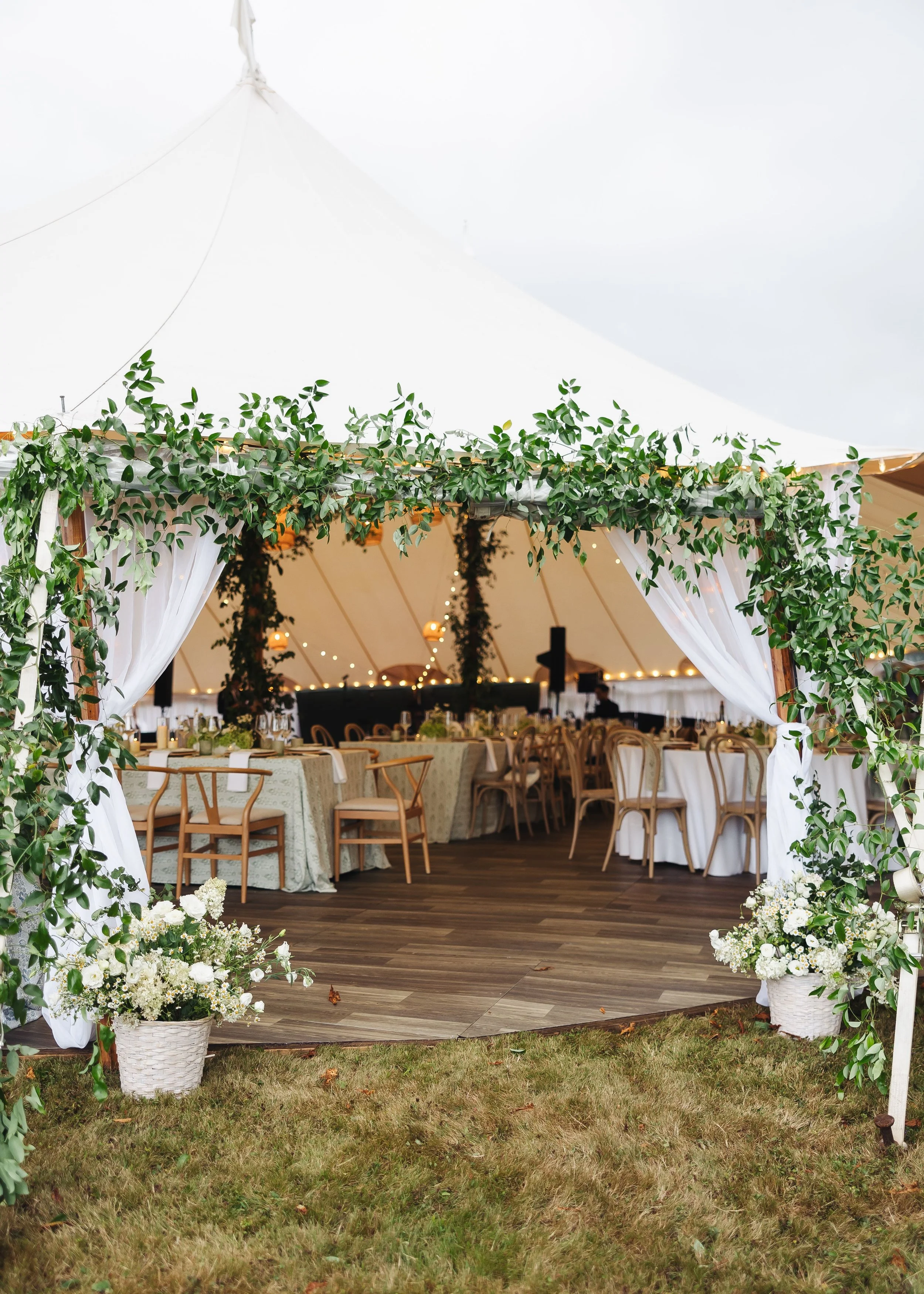 A decorated outdoor wedding reception under a large white tent with green leafy accents, white curtains, and tables with floral arrangements, wooden chairs, and string lights.