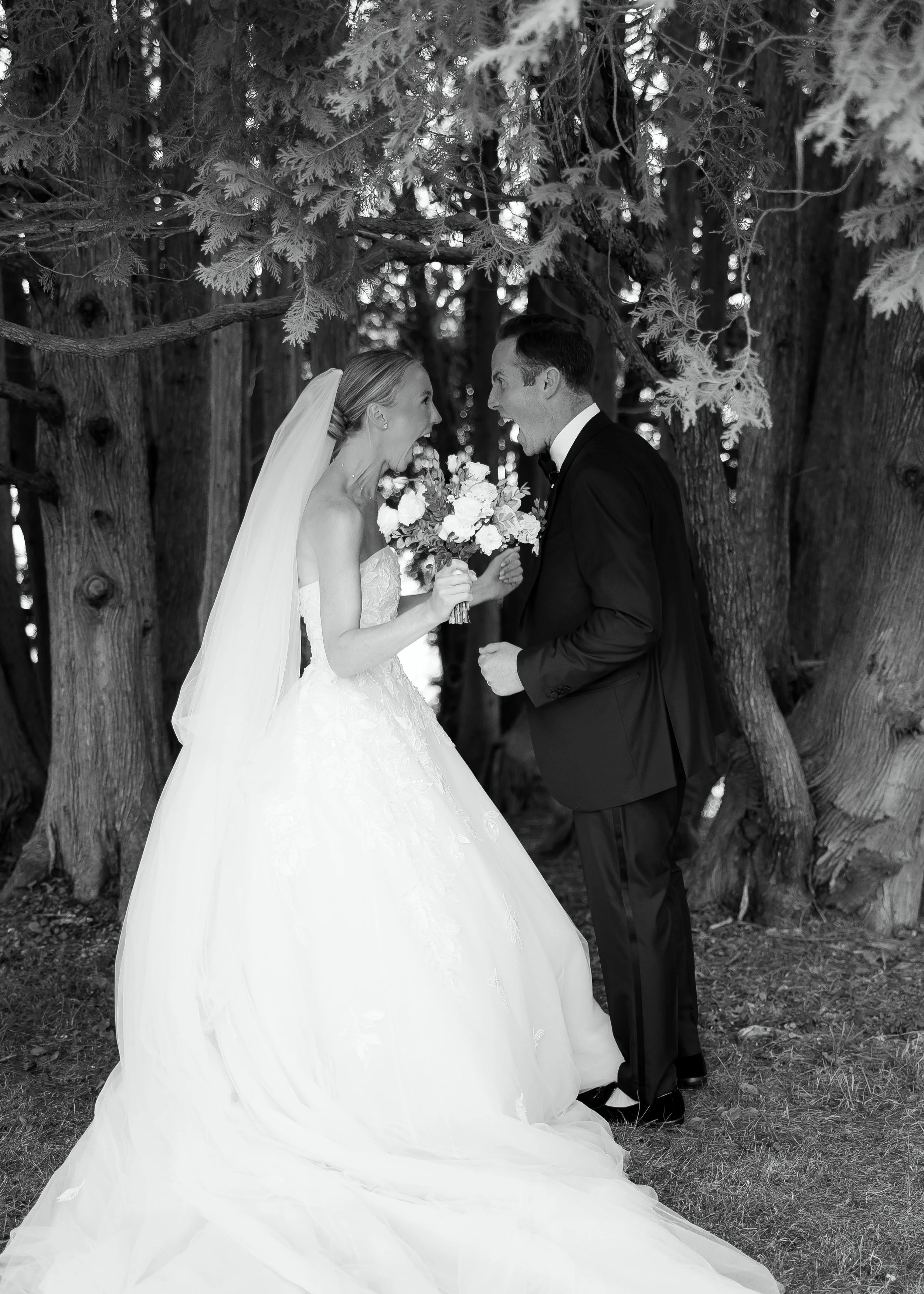 A black and white photo of a bride and groom outdoors. The bride is wearing a wedding dress with a veil, holding a bouquet, and the groom is in a tuxedo. They are smiling and leaning towards each other under a tree.
