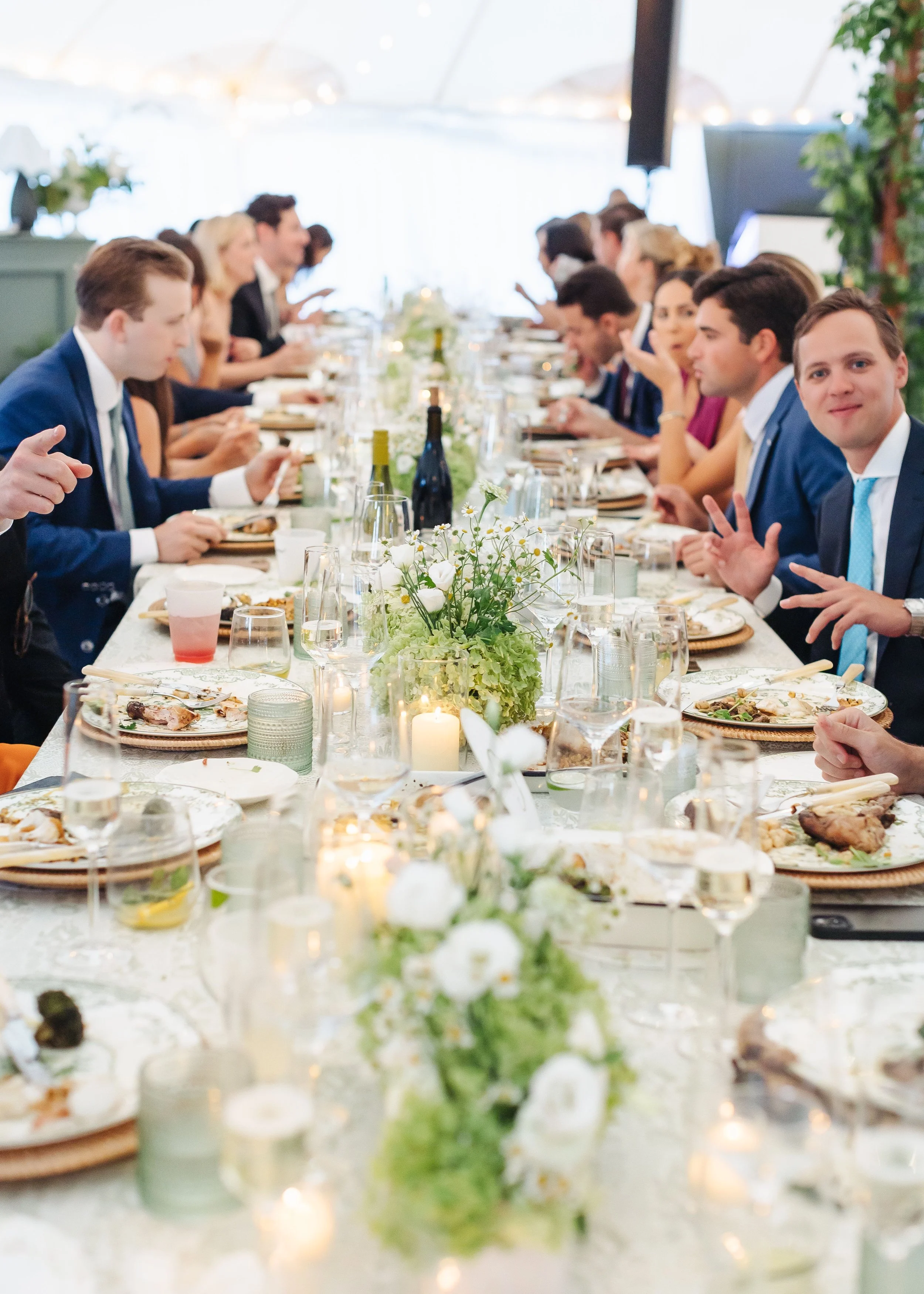 Long banquet table with white tablecloth, set for a formal gathering, decorated with a floral centerpiece and candles. People in formal attire are sitting and dining under a canopy.