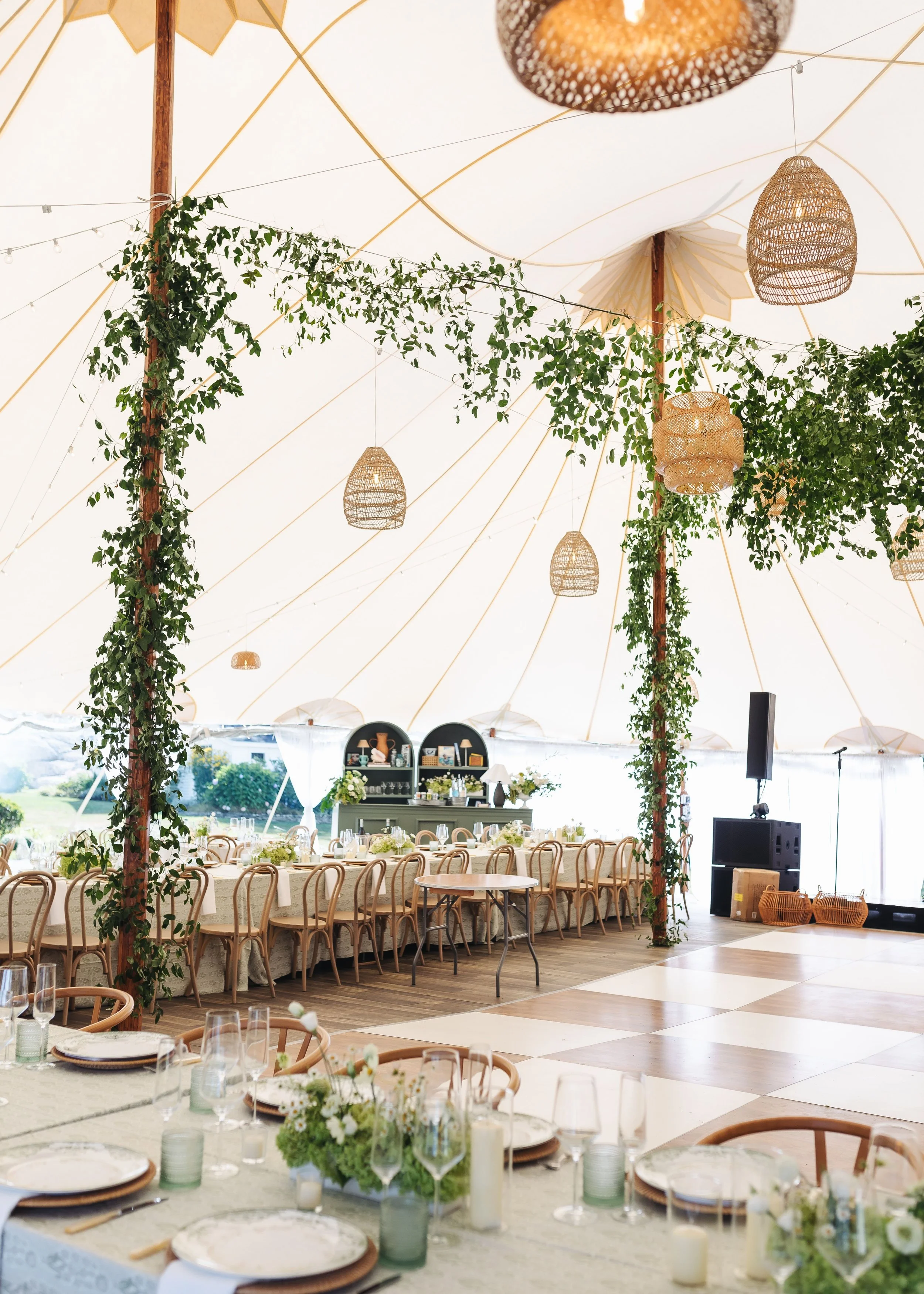 A decorated event tent with white ceiling, wooden poles, hanging wicker lamps, and green leafy garlands. Inside, long tables are set with floral centerpieces, candles, and place settings, suggesting a wedding or celebration.