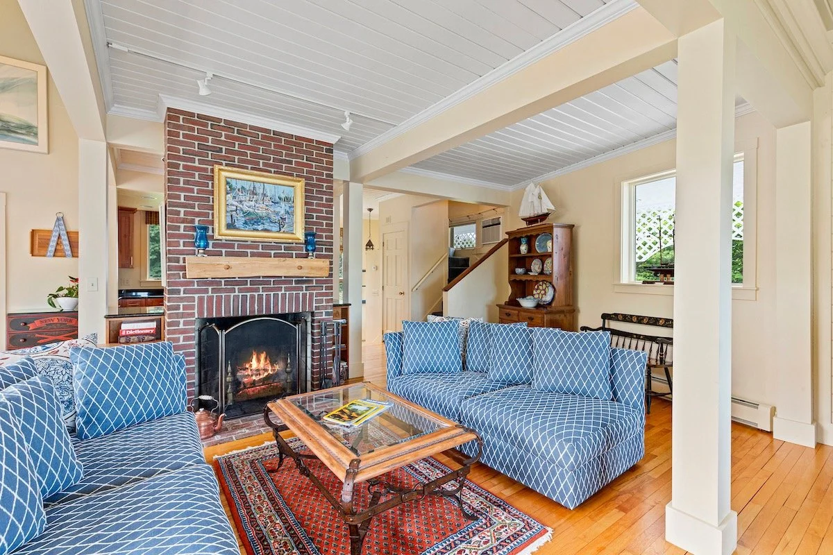 Cozy living room with a brick fireplace, blue patterned sofas, a wooden coffee table with magazines, a wooden cabinet with plates, and windows with outdoor views.