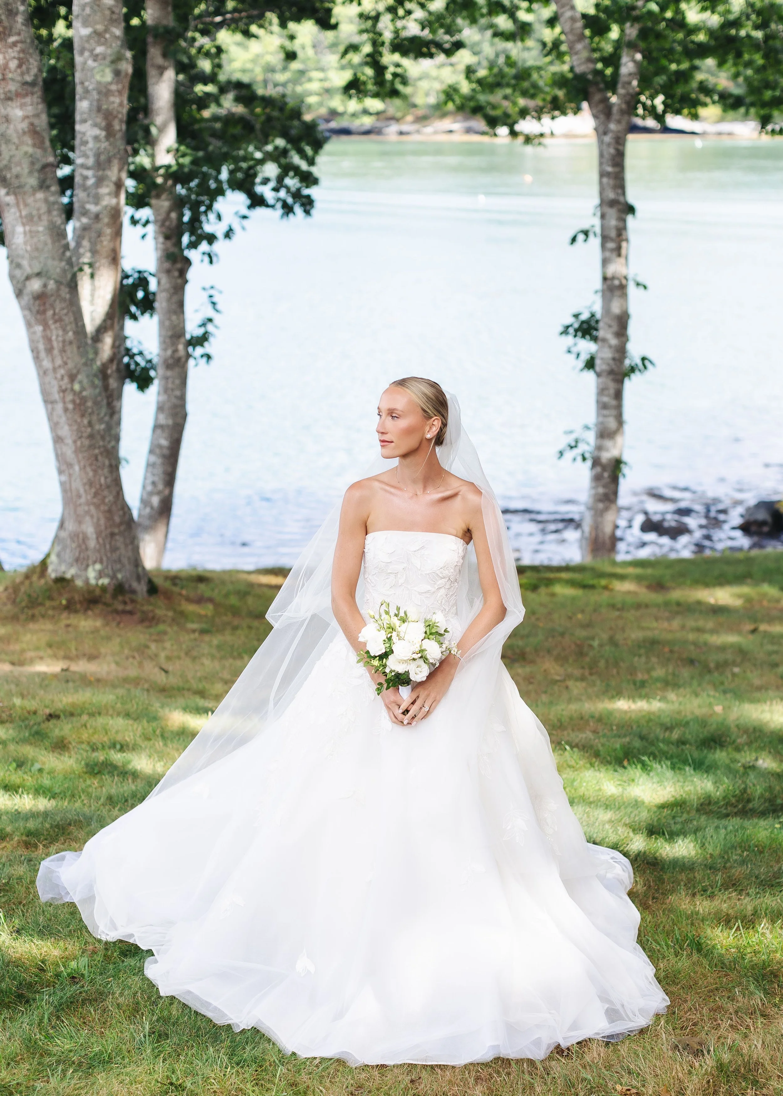 A bride in a white wedding gown holding a bouquet outdoors by a waterfront, surrounded by trees with green leaves.