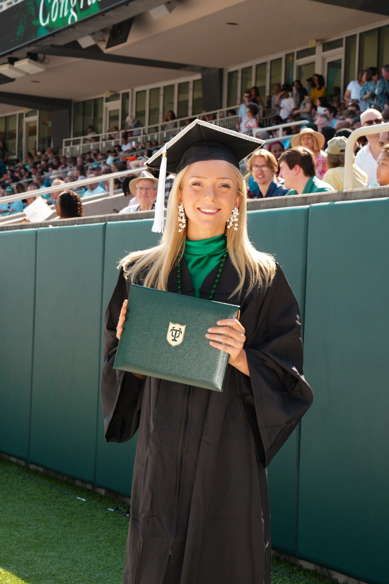 A young woman in graduation attire holding a diploma, smiling, with a large gathering of people seated in stadium bleachers behind her.