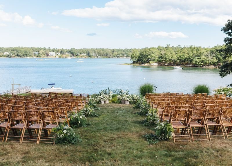 A coastal outdoor wedding ceremony site with rows of wooden chairs facing the water, decorated with white flowers and greenery, set on a grassy area.